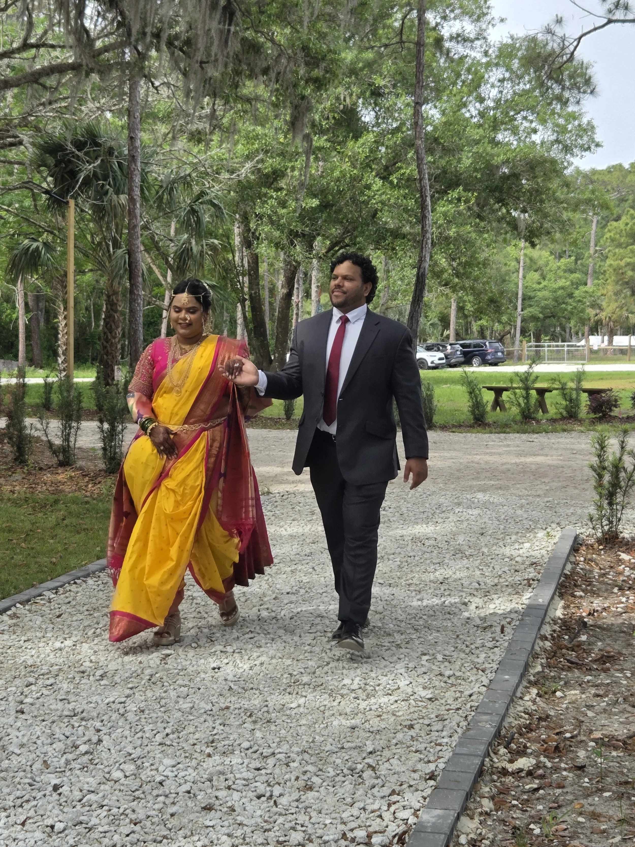 A man and a woman walking outdoors on a gravel pathway surrounded by trees. The woman is wearing a colorful sari and gold jewelry, and the man is in a dark suit with a white shirt and red tie. They are holding hands.