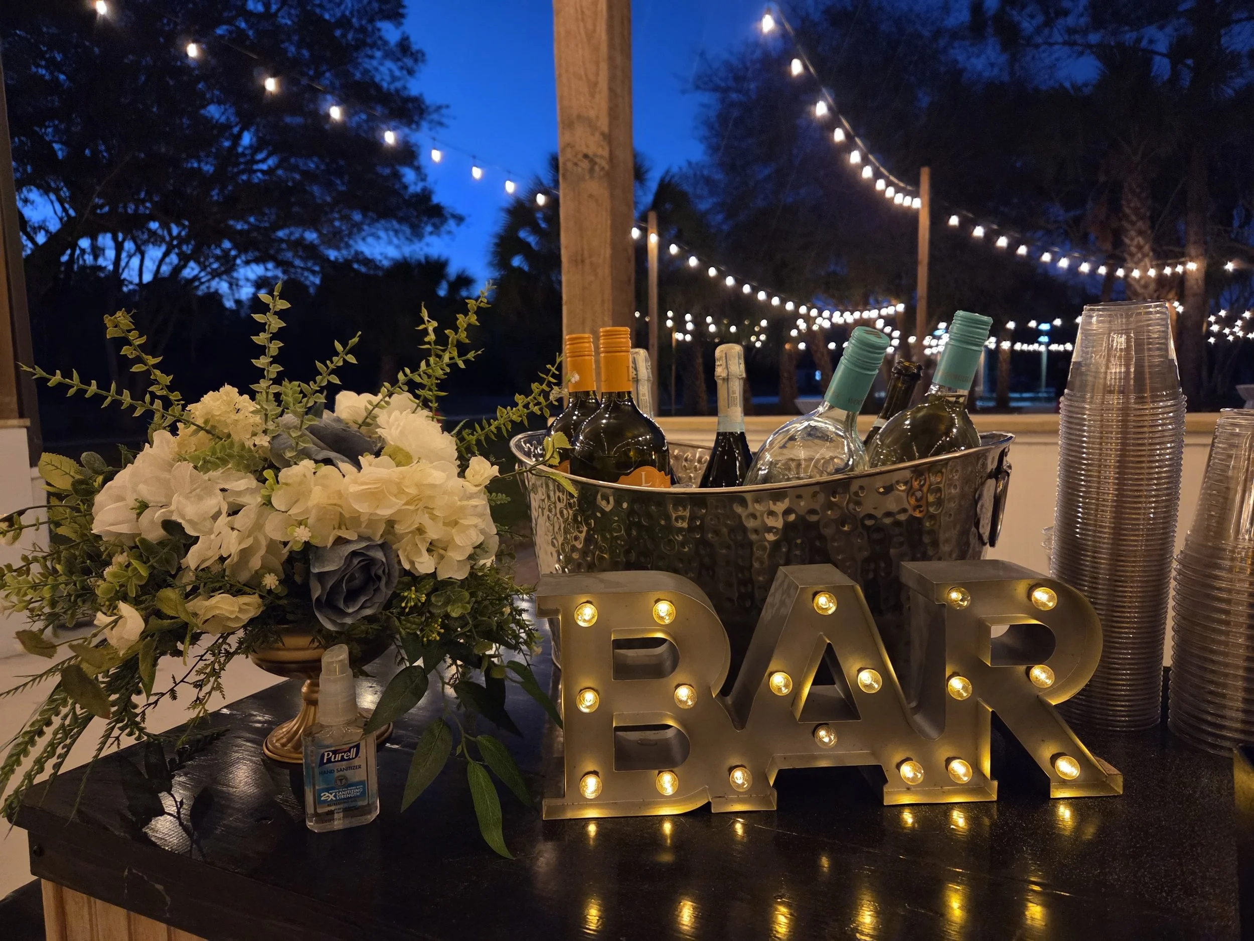 Nighttime outdoor scene with string lights, a table with wine bottles, glasses, a bouquet of flowers, and a decorative marquee sign spelling 'LOVE' with illuminated bulbs.