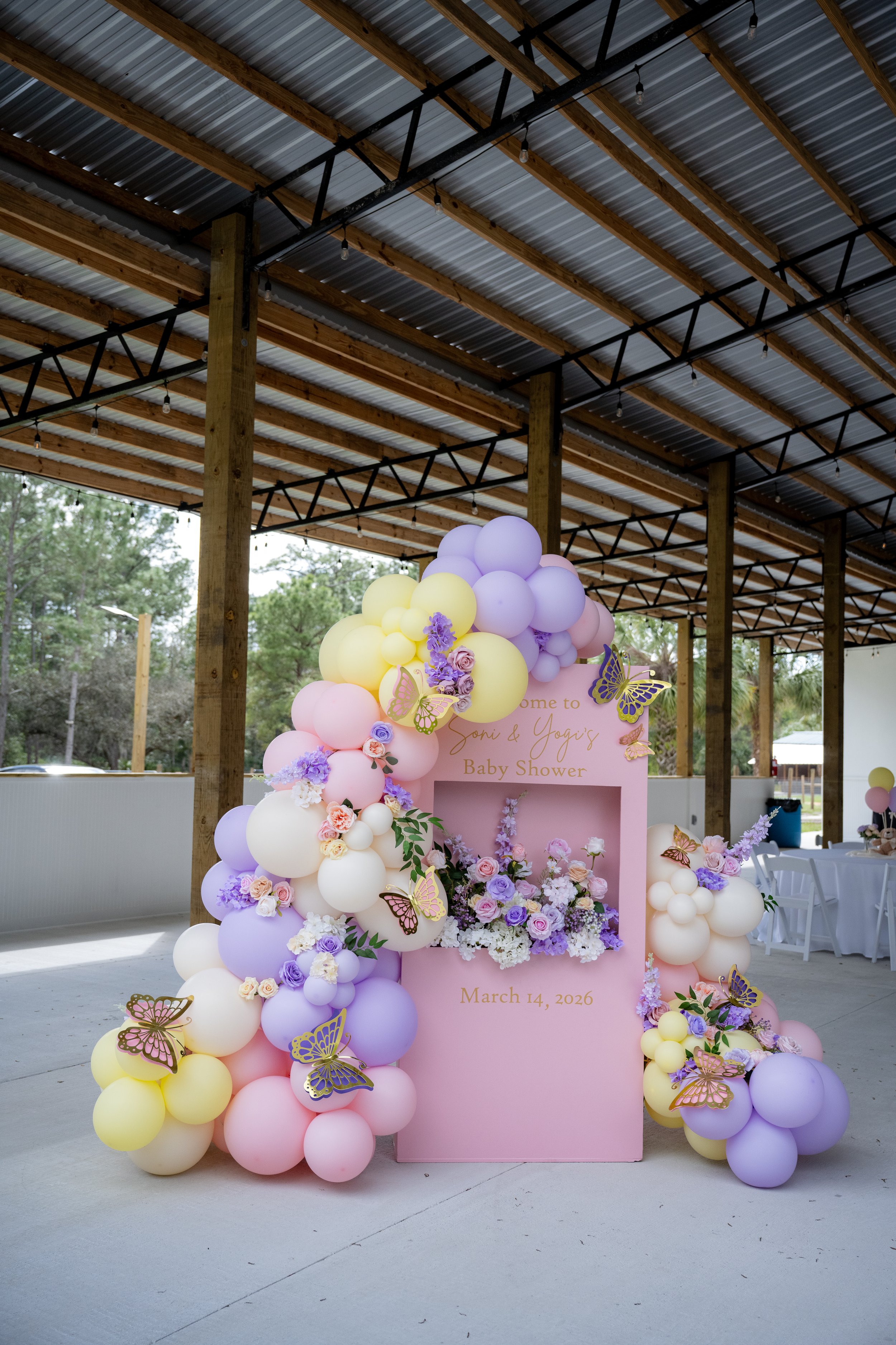 Decorative balloon arrangement for a baby shower with purple, yellow, pink, and white balloons and butterfly decorations, featuring a pink backdrop with the message 'Welcome to Soni & Yogis Baby Shower, March 14, 2026'.