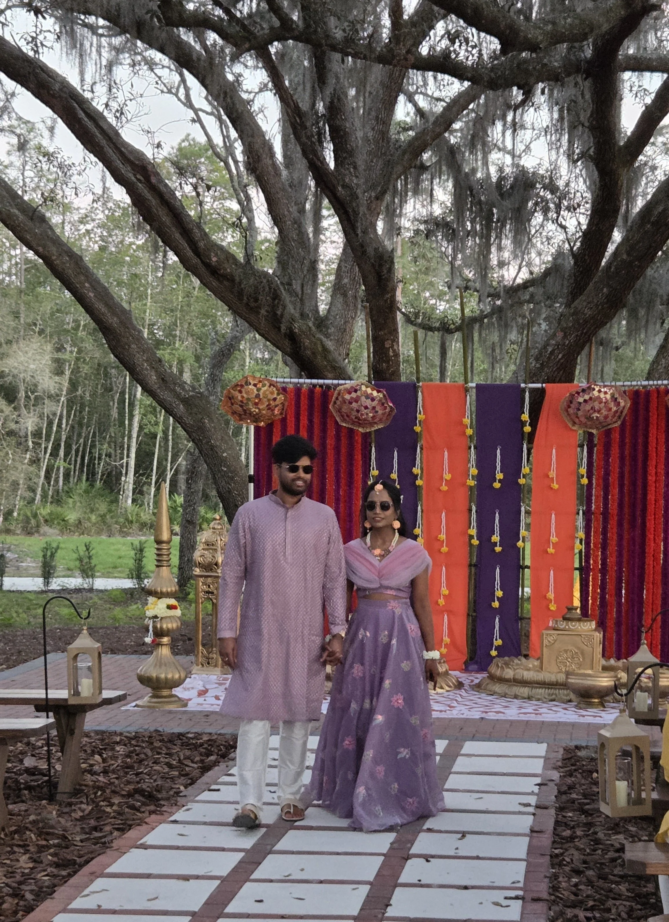A couple in traditional Indian attire holding hands, standing outdoors on a decorated walkway with lanterns, flowers, and colorful fabric backdrop, likely during a celebration or wedding.