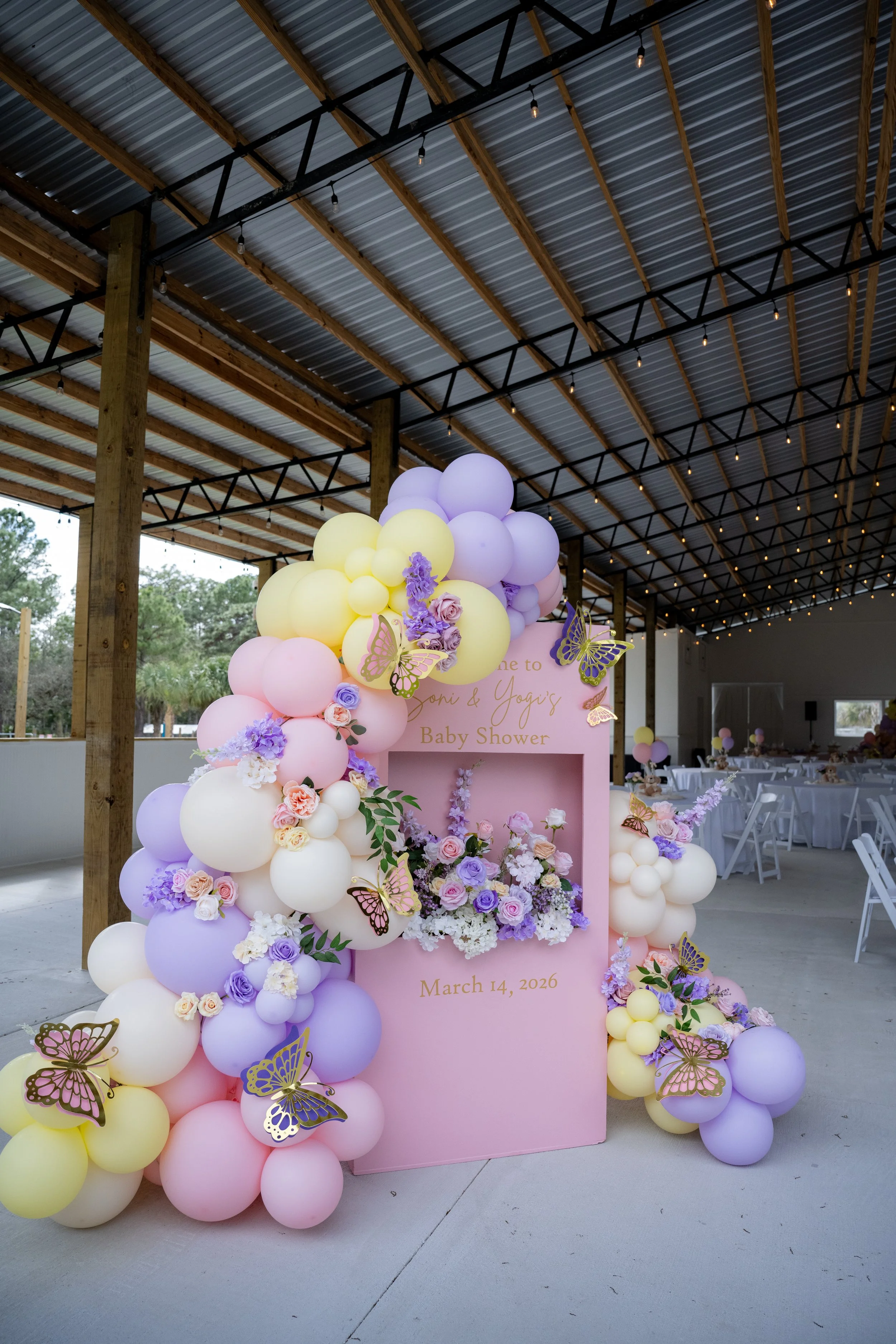 Decorative display for a baby shower with pastel-colored balloons, purple and yellow flowers, butterfly ornaments, and a pink backdrop with gold text. The event is held in a large open-sided structure with a corrugated metal roof and string lights.