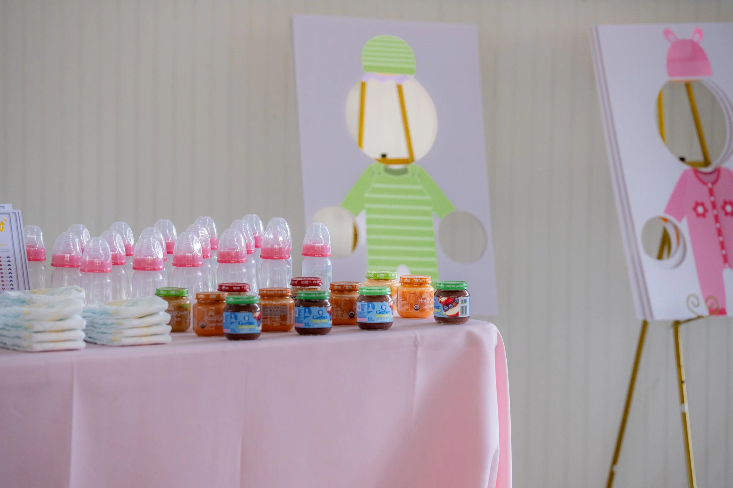 A table covered with a pink tablecloth displaying baby food in jars, baby bottles, and diapers, with colorful cartoon posters of baby characters on the wall behind.