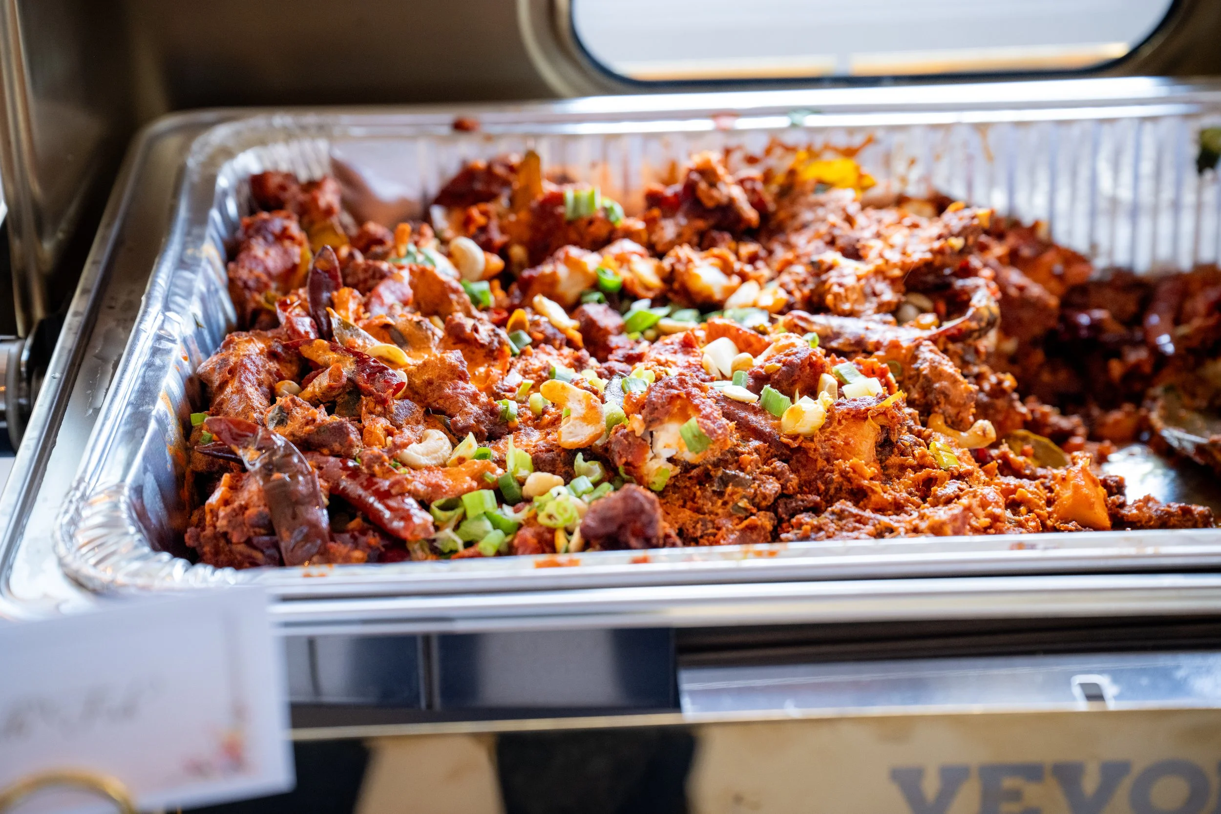 Aluminum tray of barbecue pulled chicken topped with chopped green onions, inside a cooler or heated display case.