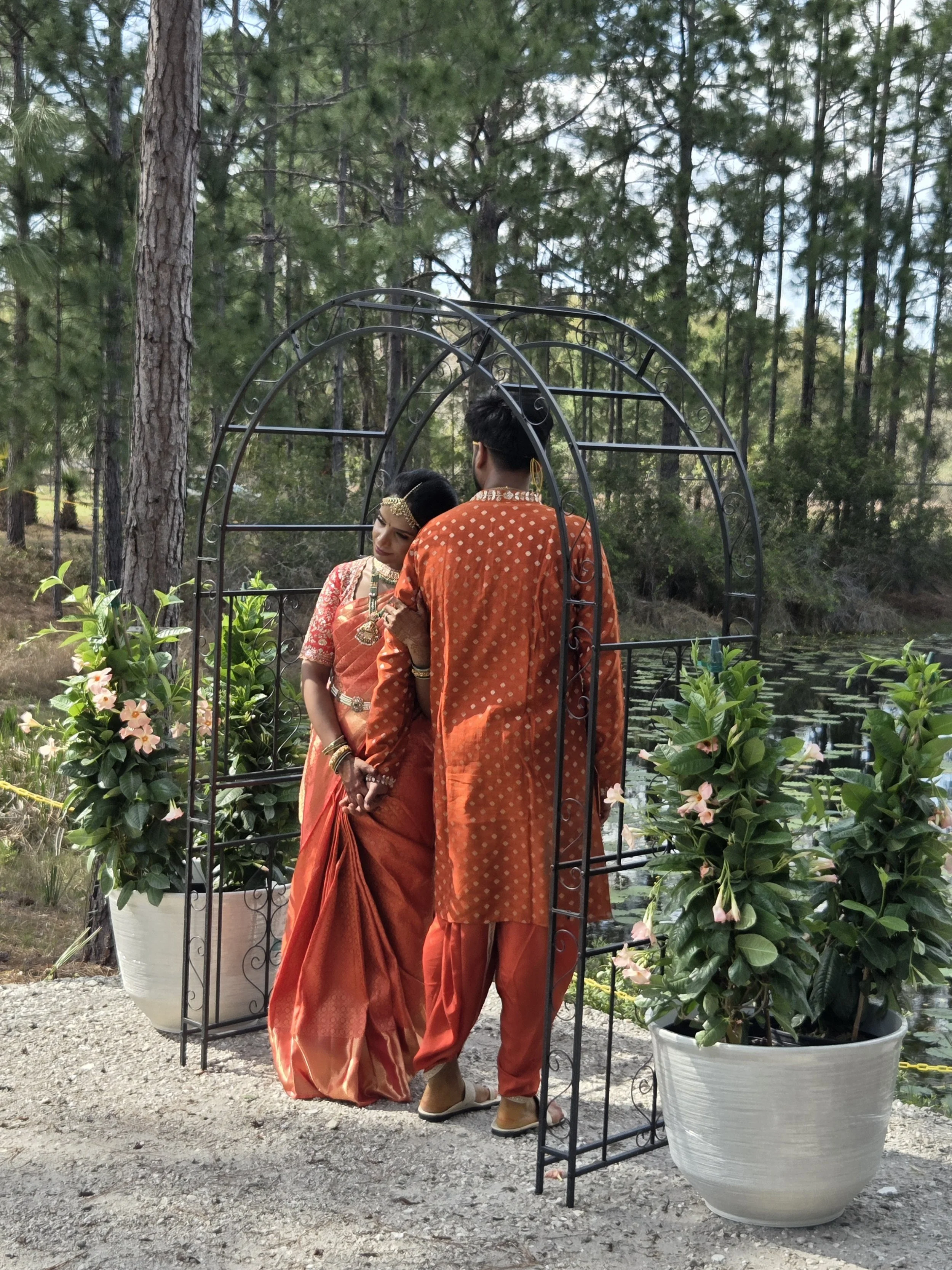 A couple dressed in traditional Indian clothing standing under a decorative metal archway near water lilies and surrounded by trees.