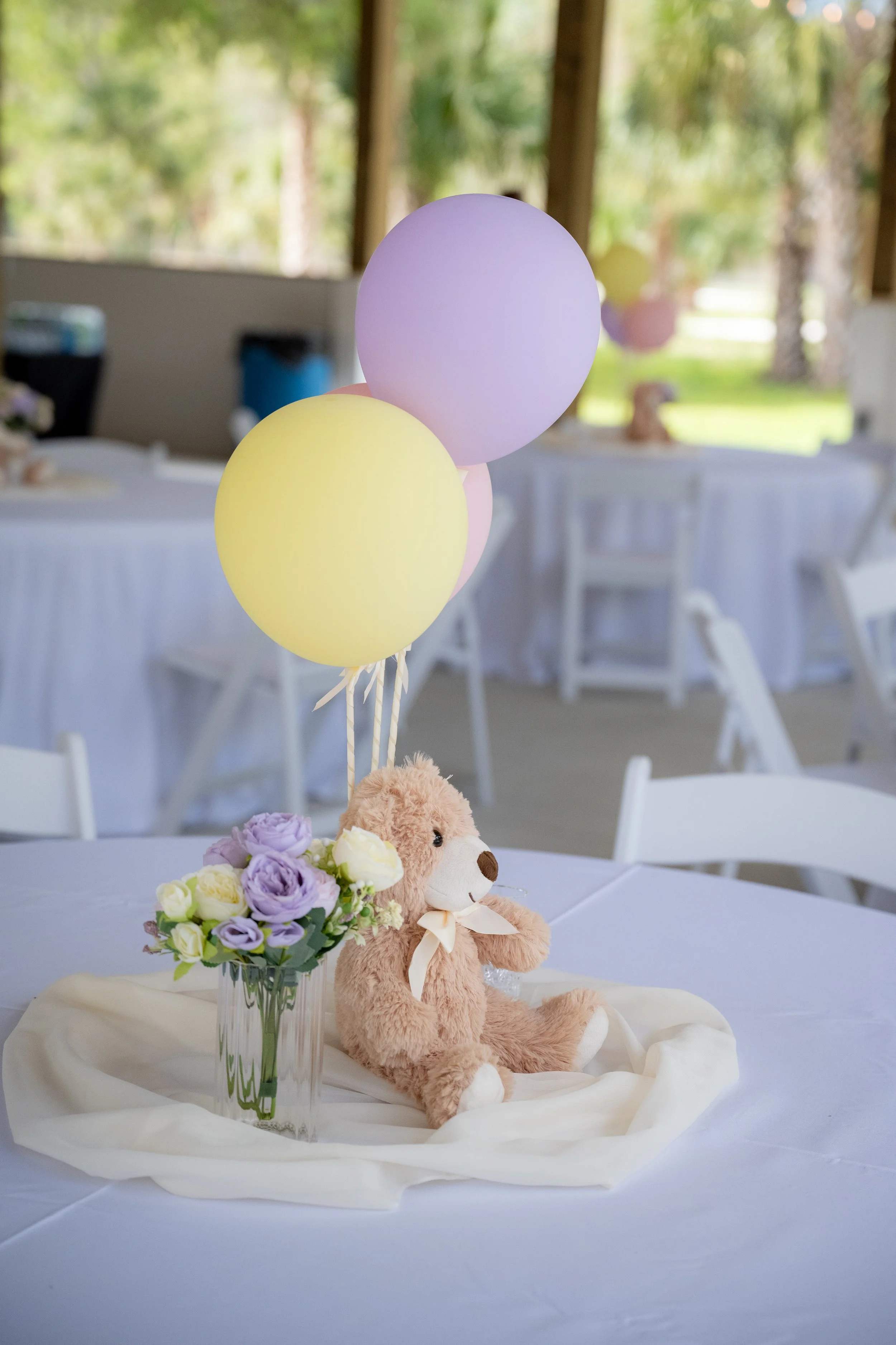 Decorated table with a bouquet of colored flowers, a plush teddy bear with a ribbon, and pastel-colored balloons in a bright indoor setting.