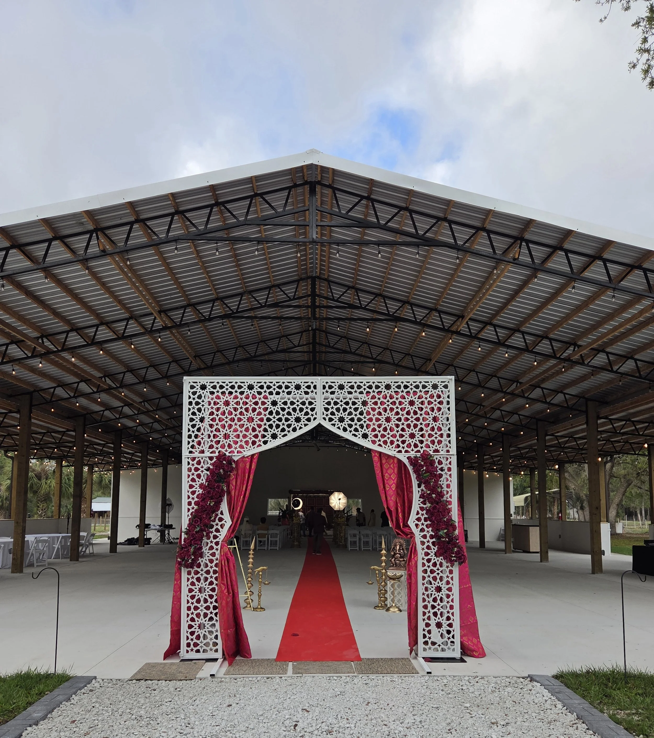 A wedding arch decorated with pink flowers and pink drapes, red carpet leading up to it, with chairs and people in the background under a large open-sided structure with a metal roof and string lights, set for a wedding ceremony.