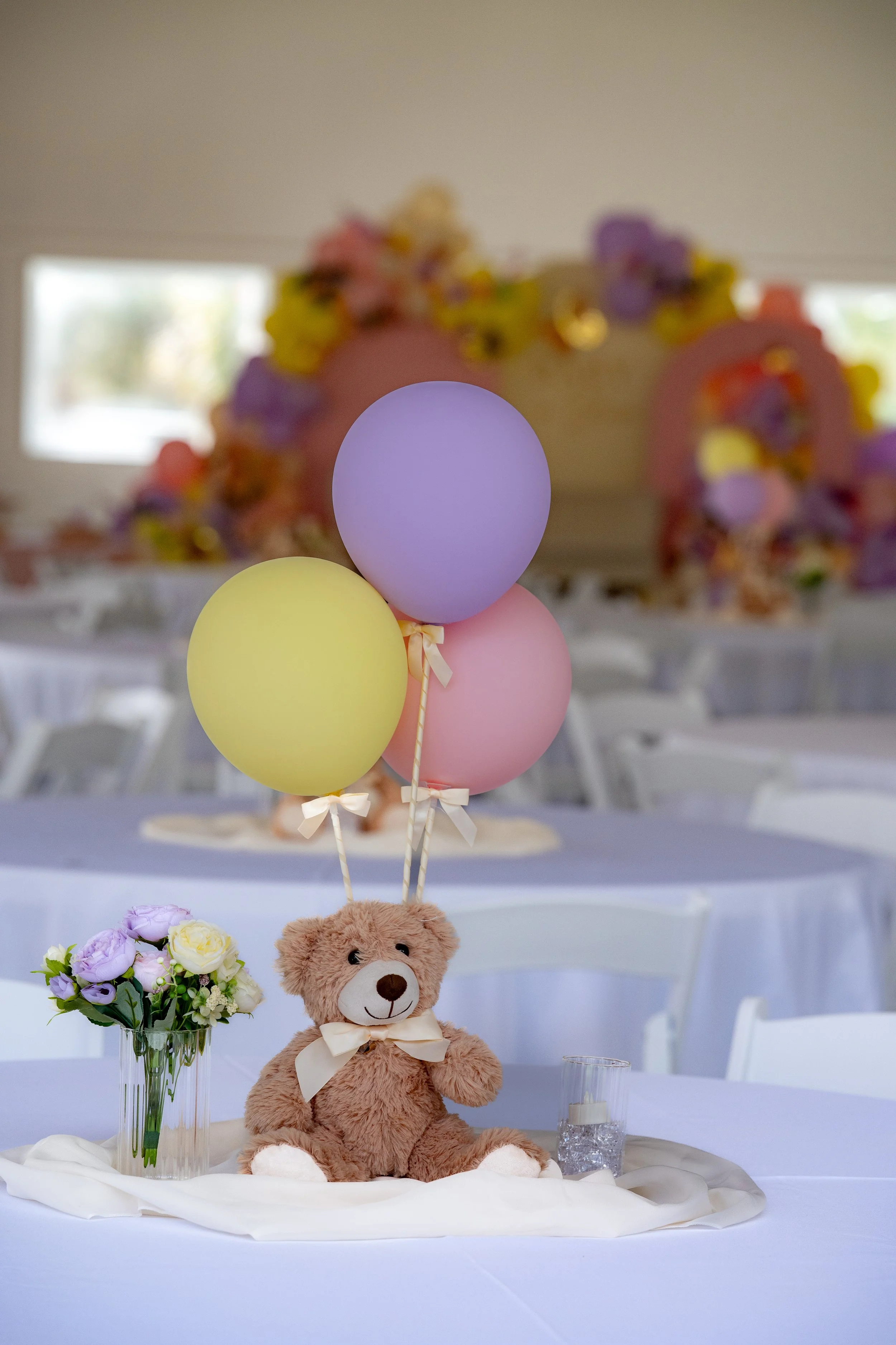 Teddy bear with a bow sitting on a table with balloons and flowers for decorations in the background.