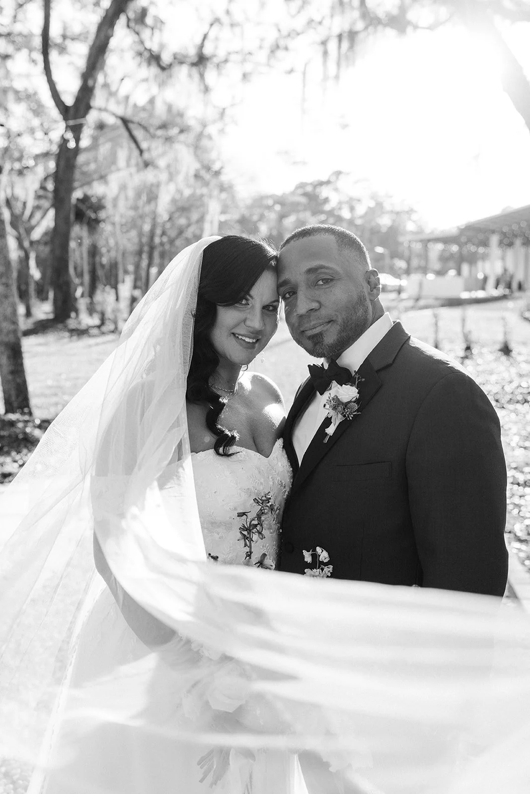 Black and white photo of a bride and groom on their wedding day outdoors, standing close with sunny background and trees.
