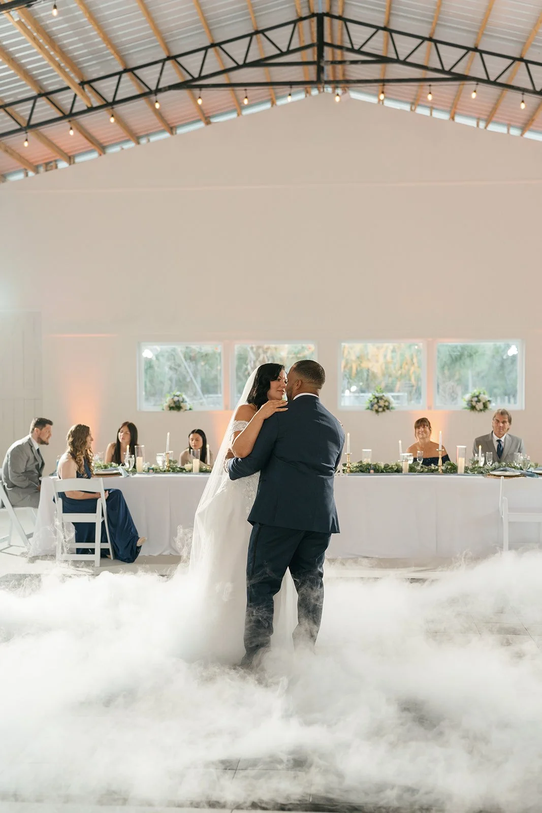 Couple dancing at wedding reception with bride and groom on the dance floor, surrounded by wedding guests seated at a long decorated table, inside a rustic venue with high ceiling and string lights.