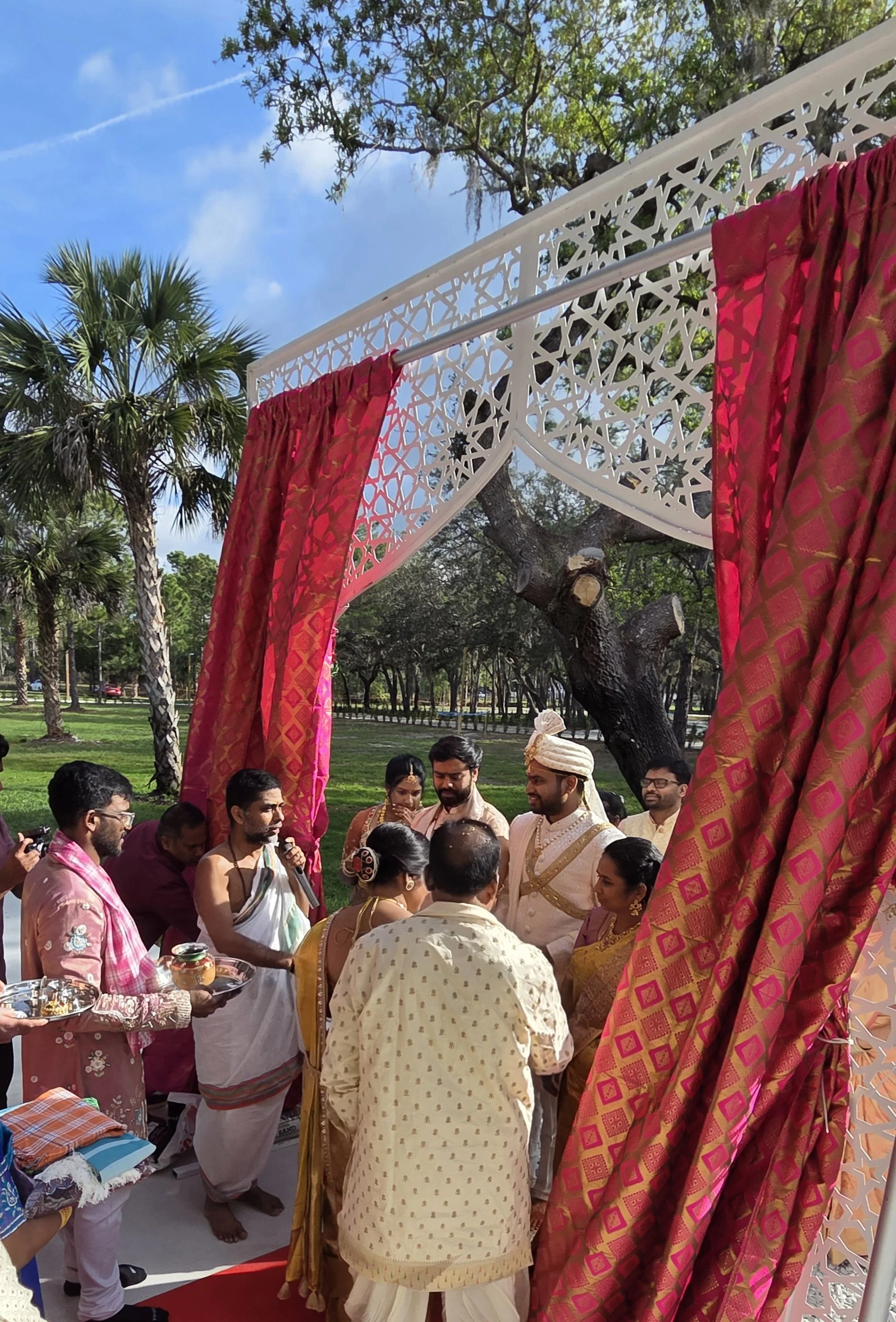 Indian wedding ceremony outdoors with people dressed in traditional attire gathered under a decorated canopy with red and gold fabrics, surrounded by trees and blue sky.
