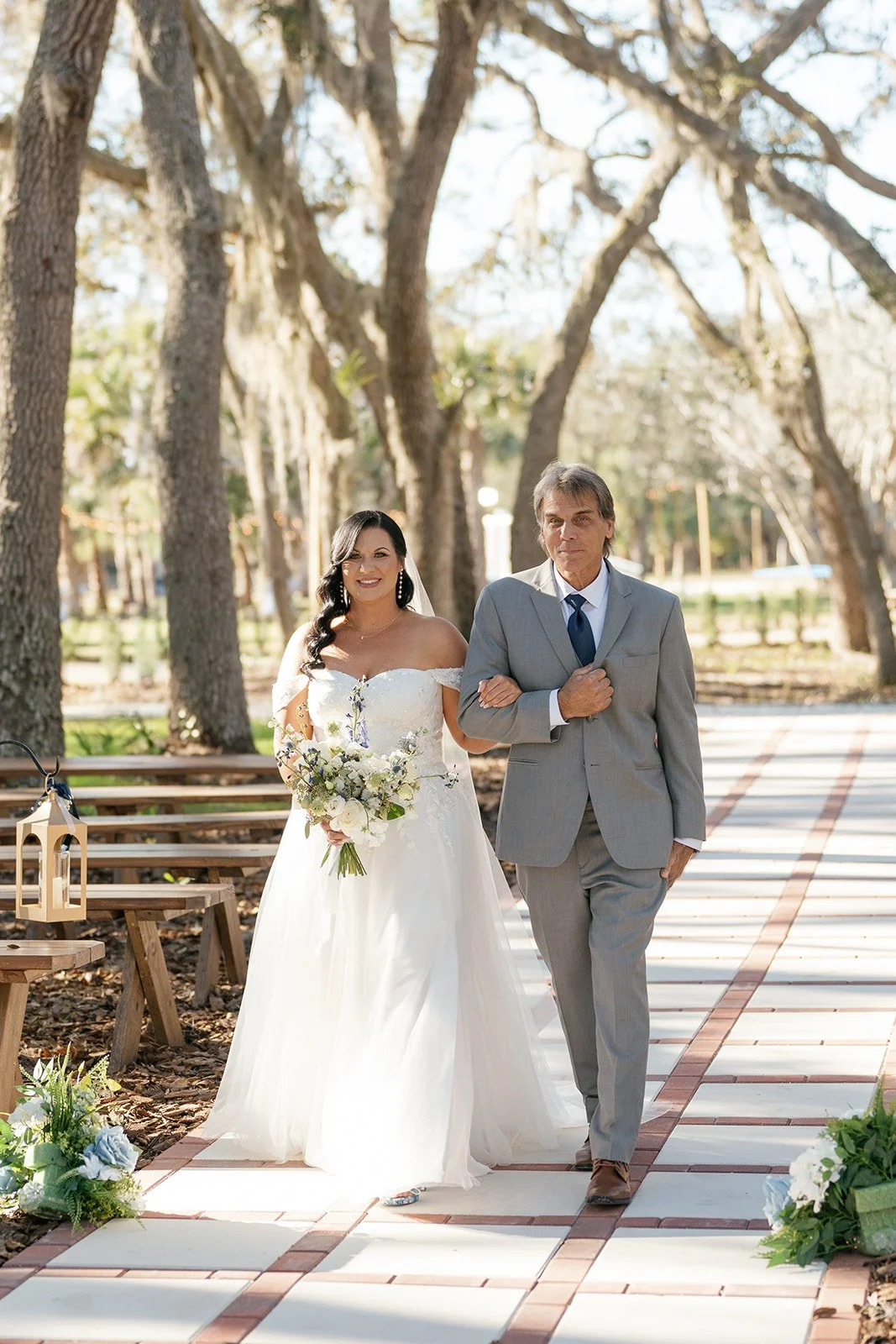A bride in a white wedding gown holding a bouquet walks arm-in-arm with a man in a gray suit on a pathway surrounded by trees and wooden benches.