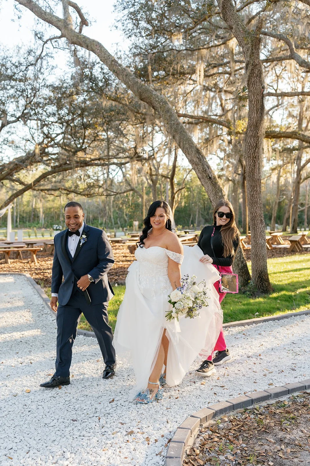 A bride in a white wedding dress walking outdoors with two people in a park-like setting with trees and benches, during daytime.