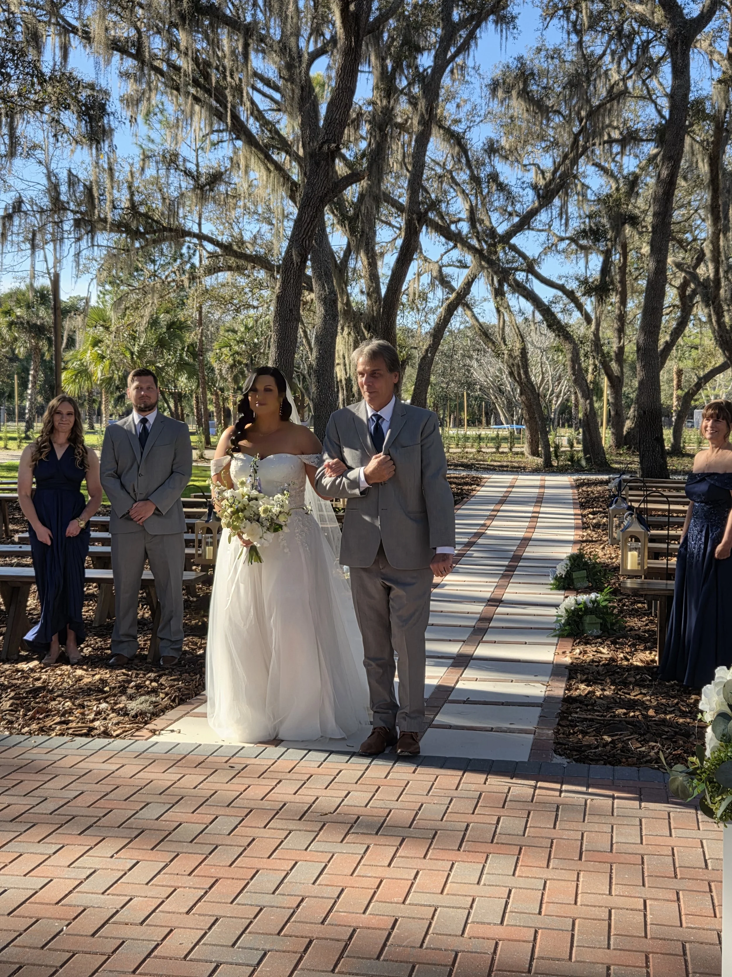 A wedding ceremony outdoors with a bride in a white dress holding a bouquet, walking with a man in a gray suit, followed by bridesmaids and groomsmen, set among trees with Spanish moss, lanterns, and a paved walkway.