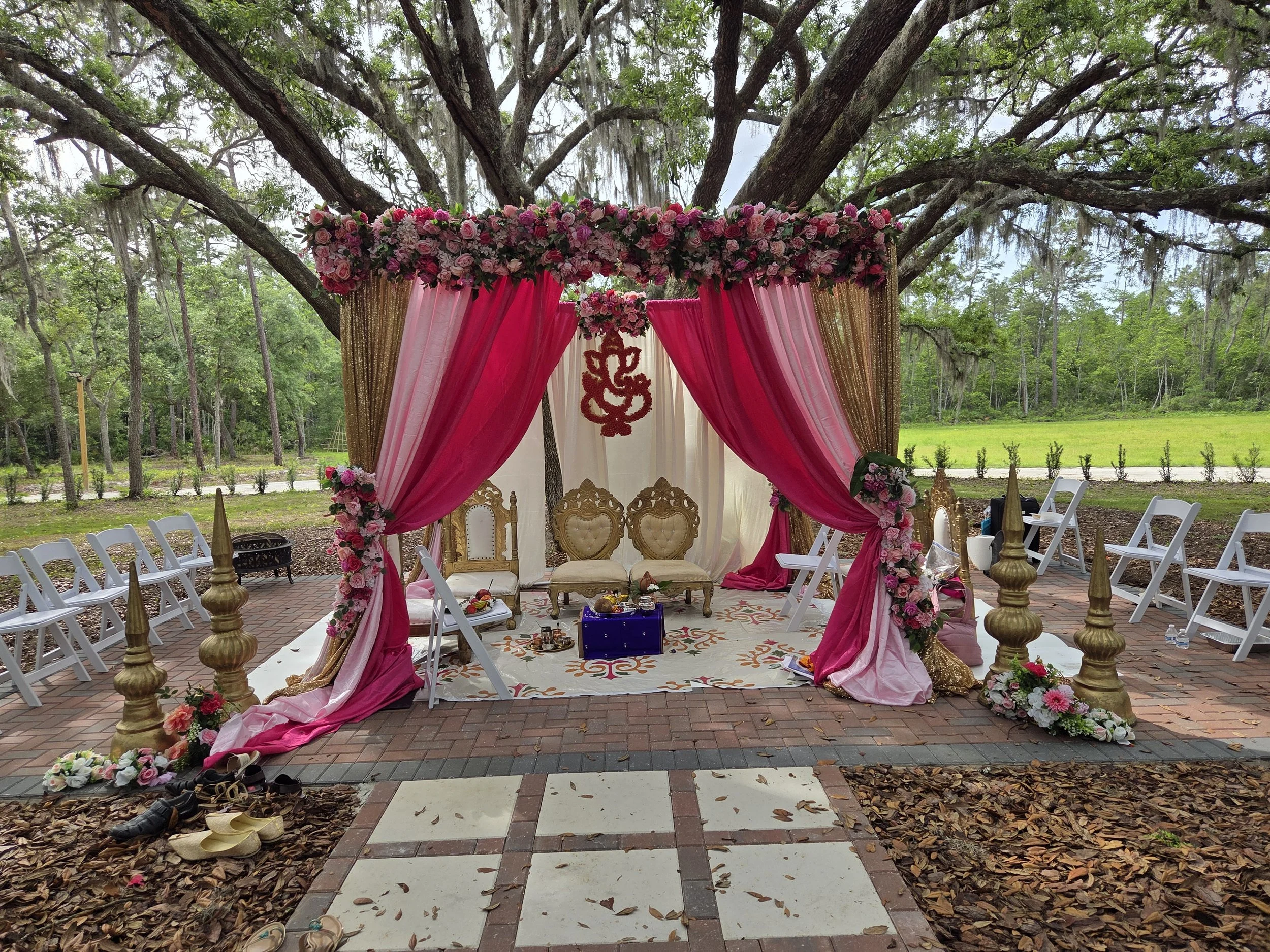 A decorated outdoor wedding altar with pink and gold drapes, floral arrangements, and chairs arranged for guests, set in a wooded area with trees in the background.
