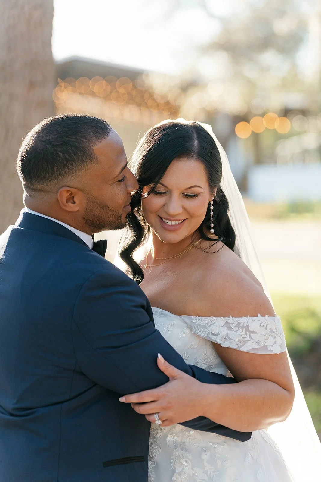A bride and groom sharing an intimate moment outdoors, with the groom in a navy tuxedo and the bride in an off-the-shoulder lace wedding dress, smiling.