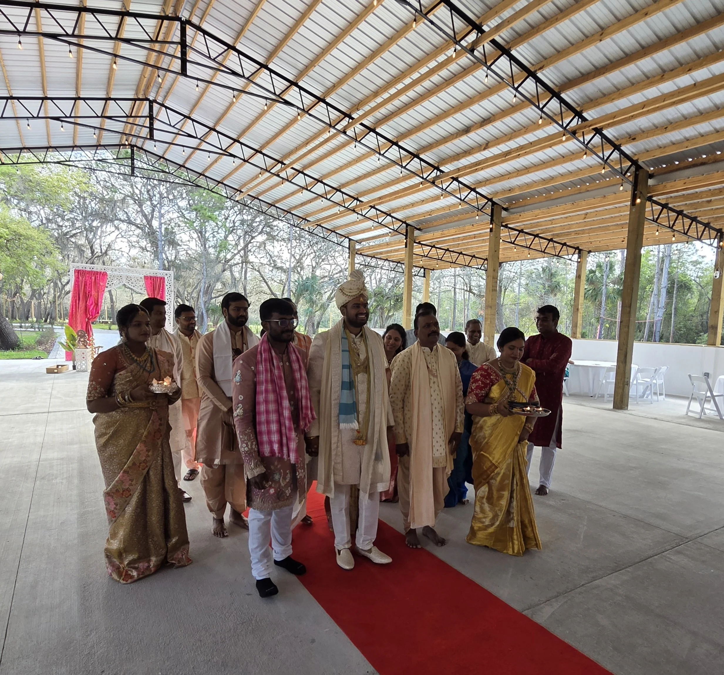 Group of people in traditional Indian attire standing on a red carpet under a large open-air wooden and metal roof structure, possibly at a festive or wedding celebration.