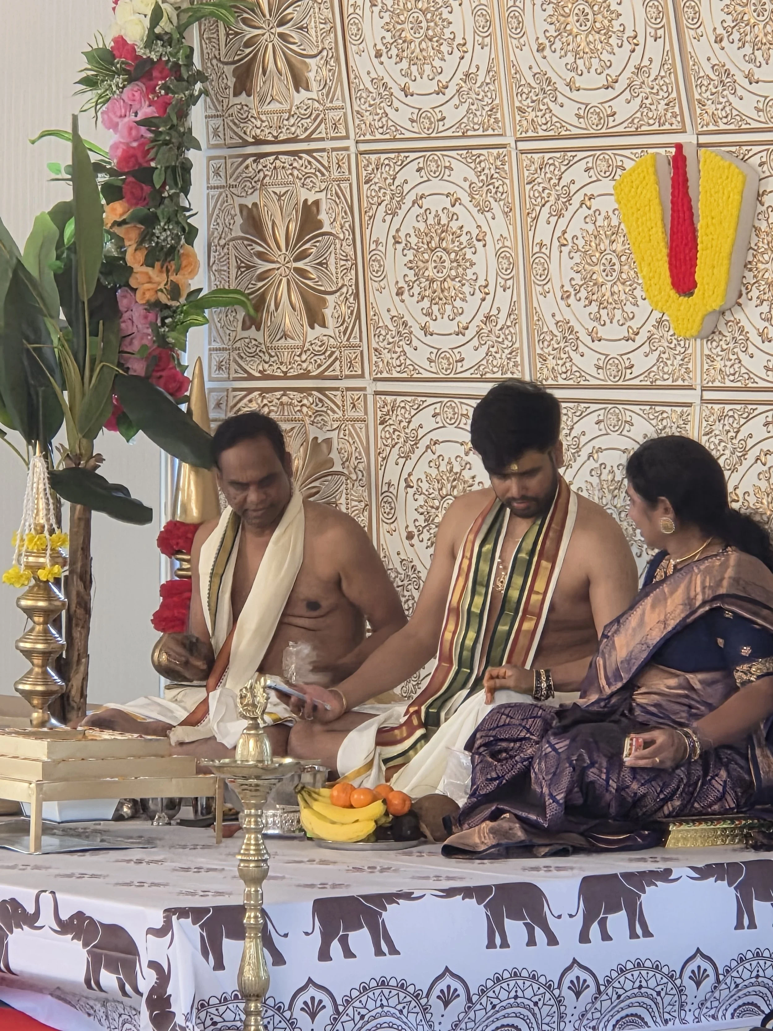 Hindu priests conducting a religious ceremony indoors, seated at a decorated altar with flowers, fruits, and religious artifacts, with ornate wall panels and a floral arrangement in the background.