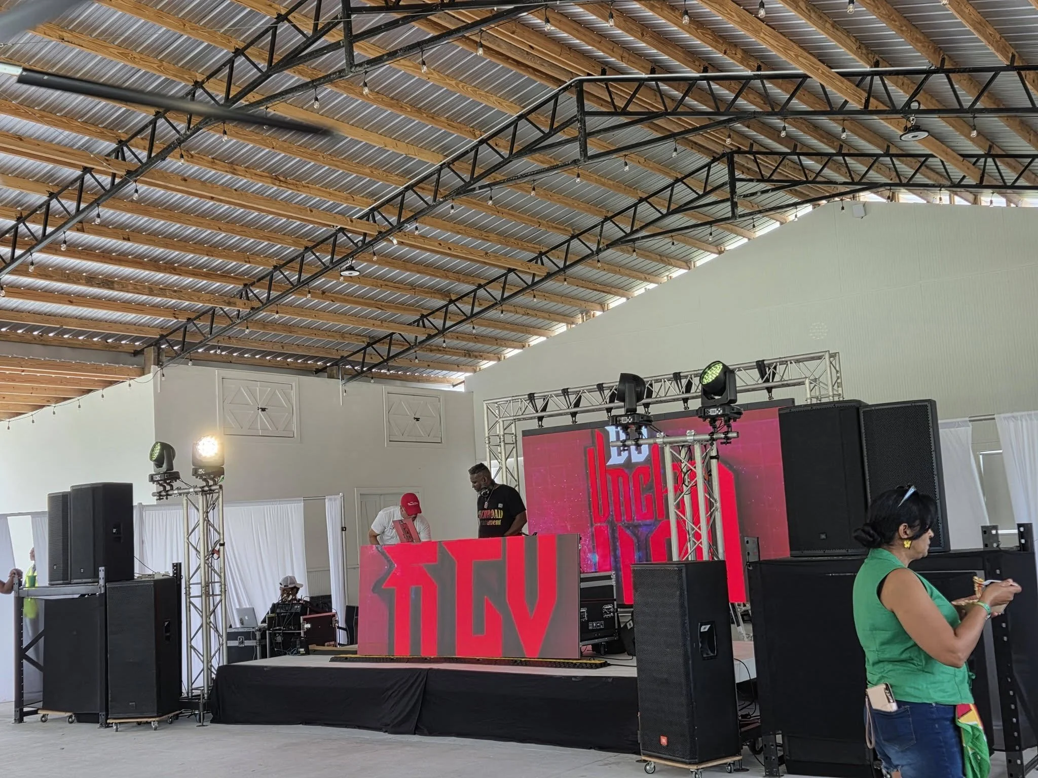 An indoor event space with a stage setup that includes DJ equipment, large speakers, and lighting. The ceiling is made of wood beams with string lights hanging down. There are a few people visible, including a woman in a green top, and a person wearing a red cap.
