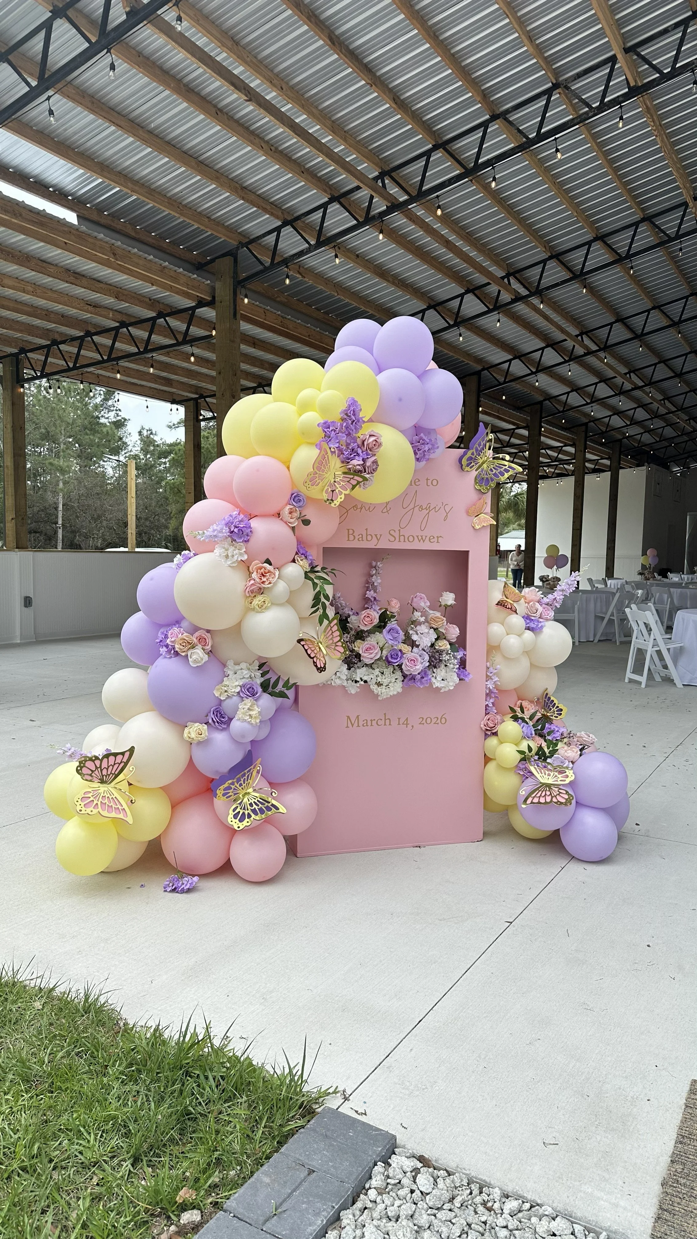 Decorative baby shower arch with pastel-colored balloons, flowers, and butterfly cutouts, in front of a pink backdrop that reads "Welcome to Toni & Yogi's Baby Shower" with date "March 14, 2026".