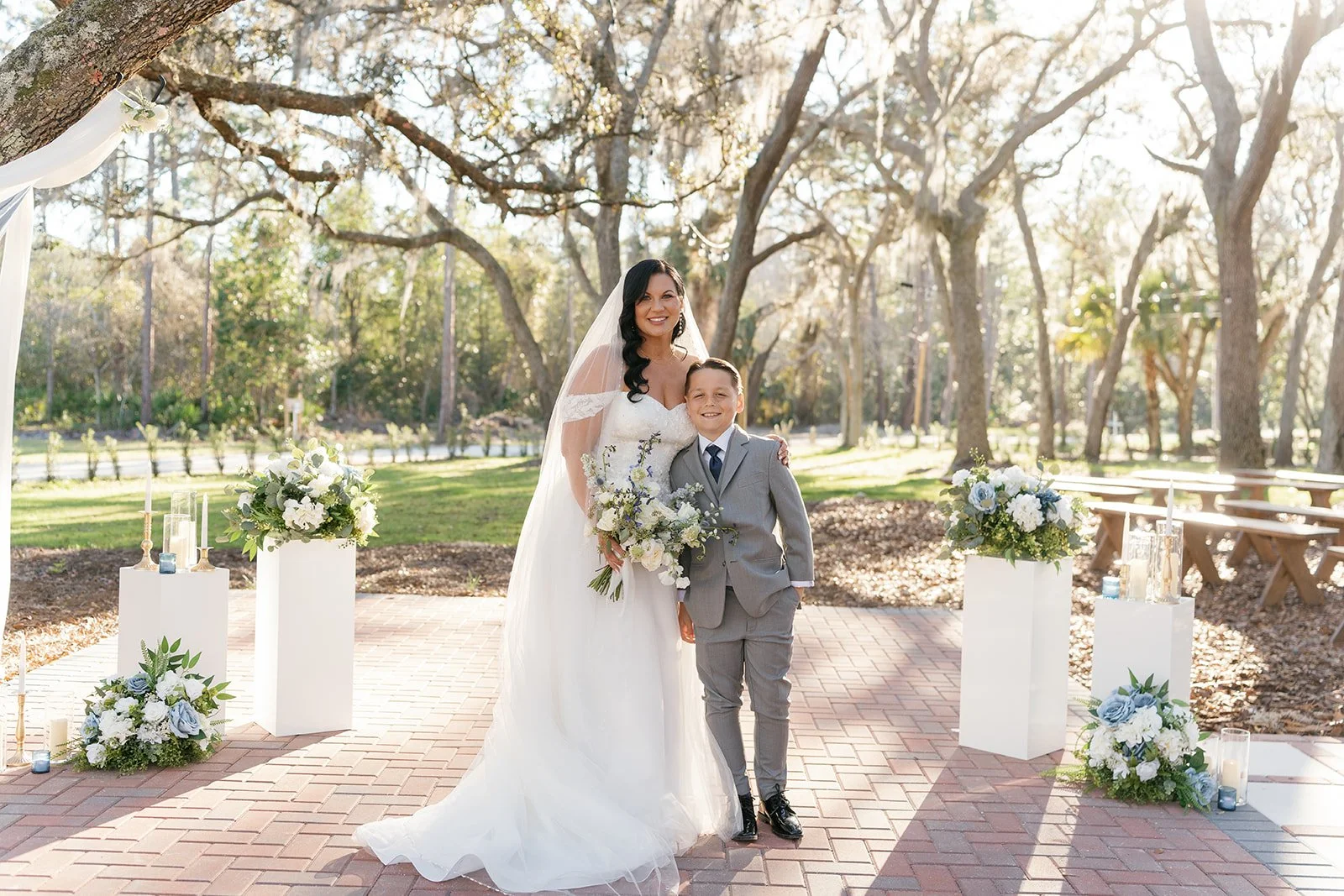 A bride and a young boy, possibly her ring bearer or a family member, smile and stand together outdoors during a wedding ceremony in a park or garden setting with trees and sunlight. The bride is wearing a white wedding gown and veil, holding a bouqu