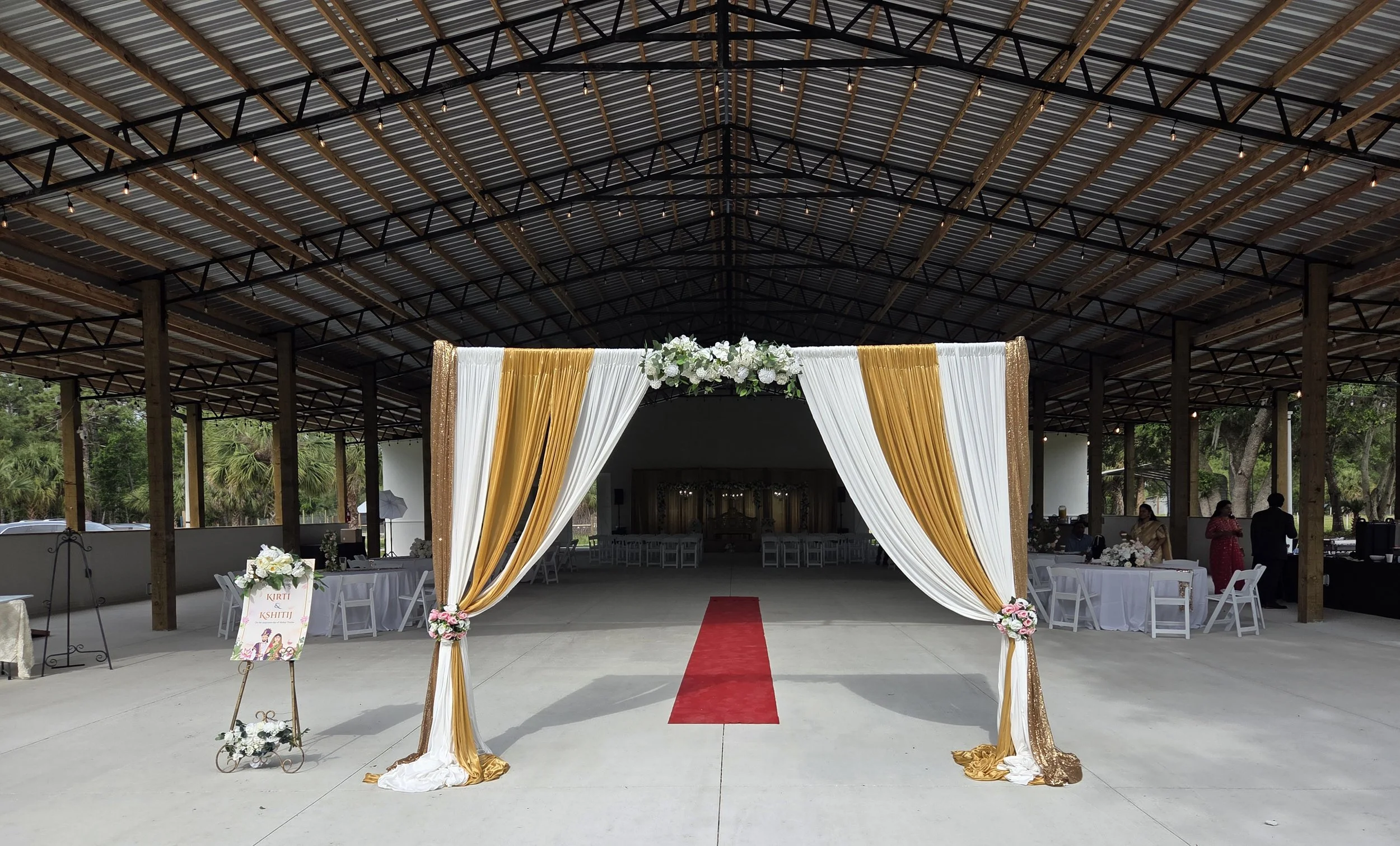 Wedding ceremony setup with a decorated arch of white and gold drapes, flowers, and a red carpet inside a large open-sided pavilion.