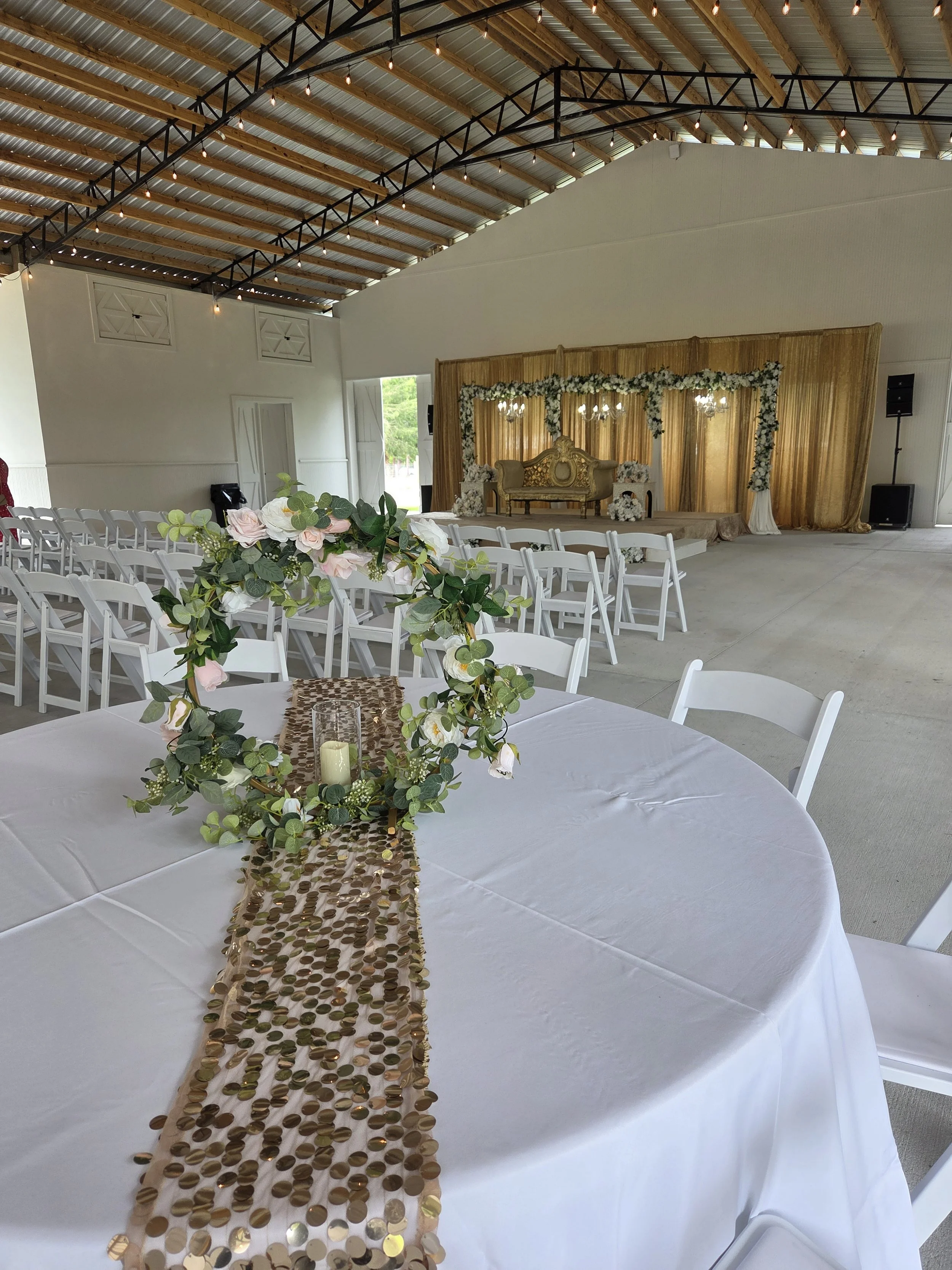 A decorated indoor event space with white chairs arranged around round tables. The central table has a white tablecloth, a gold sequin table runner, and a floral centerpiece with greenery and pink and white flowers. In the background, there is a stag