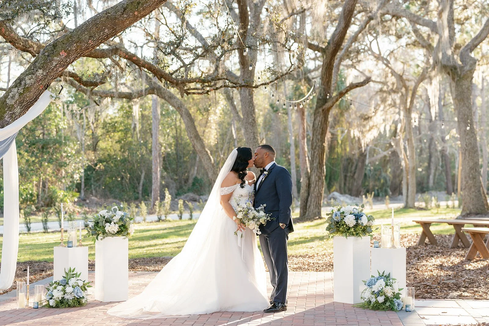 A bride and groom share a kiss during their outdoor wedding ceremony, with trees and sunlight in the background and decorated with floral arrangements and candles.