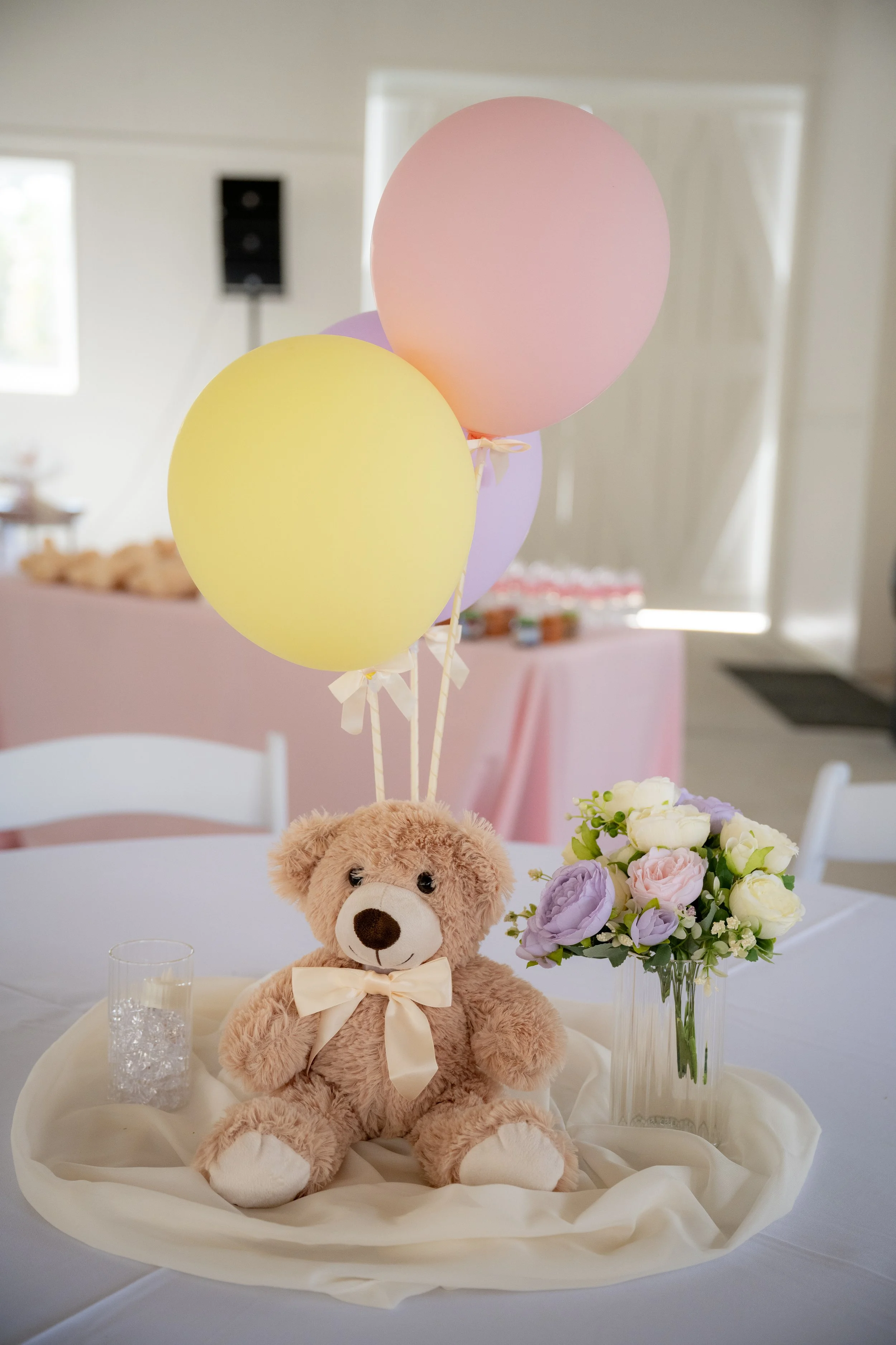A teddy bear with a cream-colored bow tie sitting on a table decorated for a celebration, with a bouquet of purple, white, and cream flowers in a tall glass vase, pastel balloons floating above, and a pink tablecloth in the background.