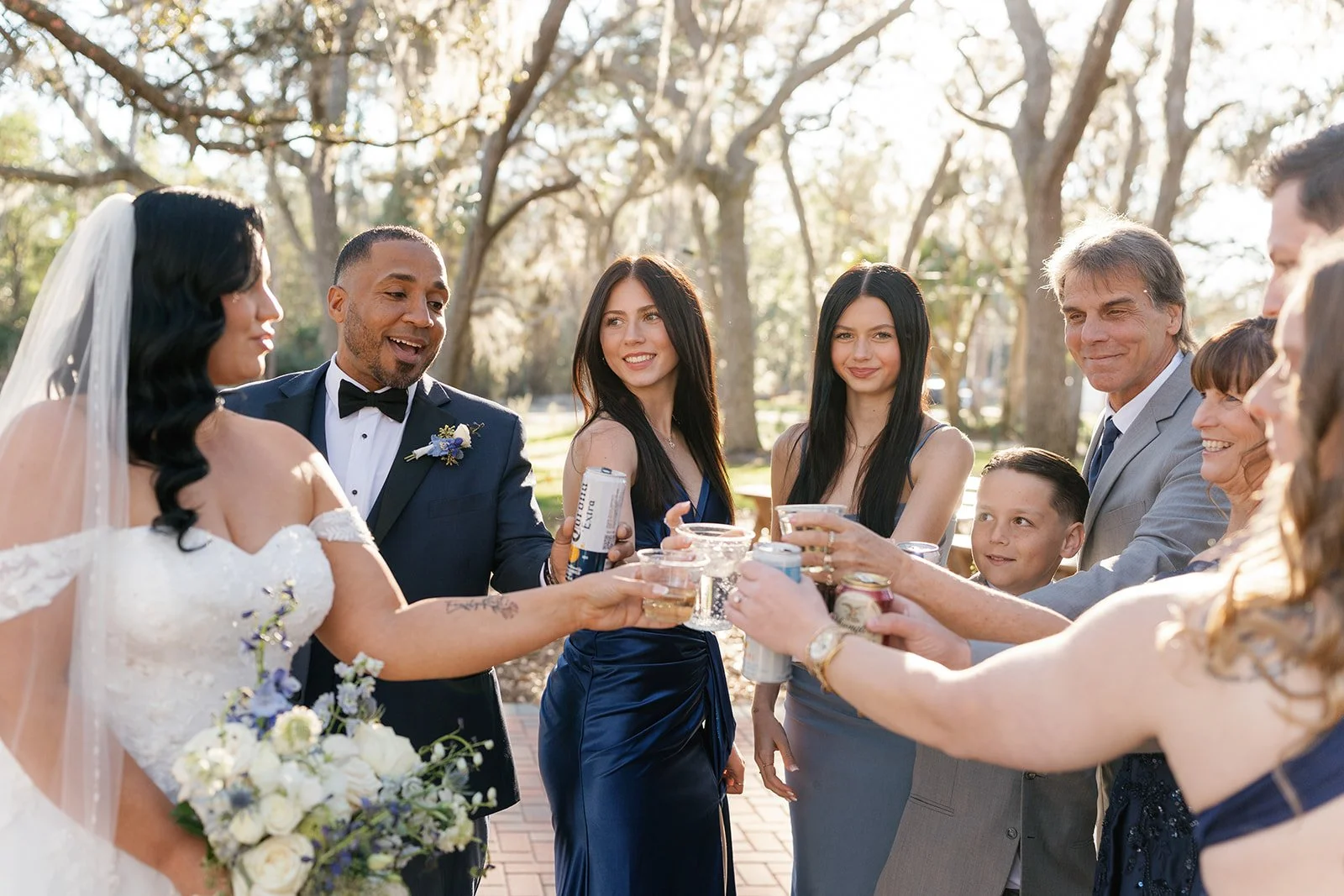 A group of people at an outdoor wedding celebration holding drinks together in a toast, with the bride in a white wedding dress and the groom in a tuxedo. The scene is set in a sunny park with trees in the background.
