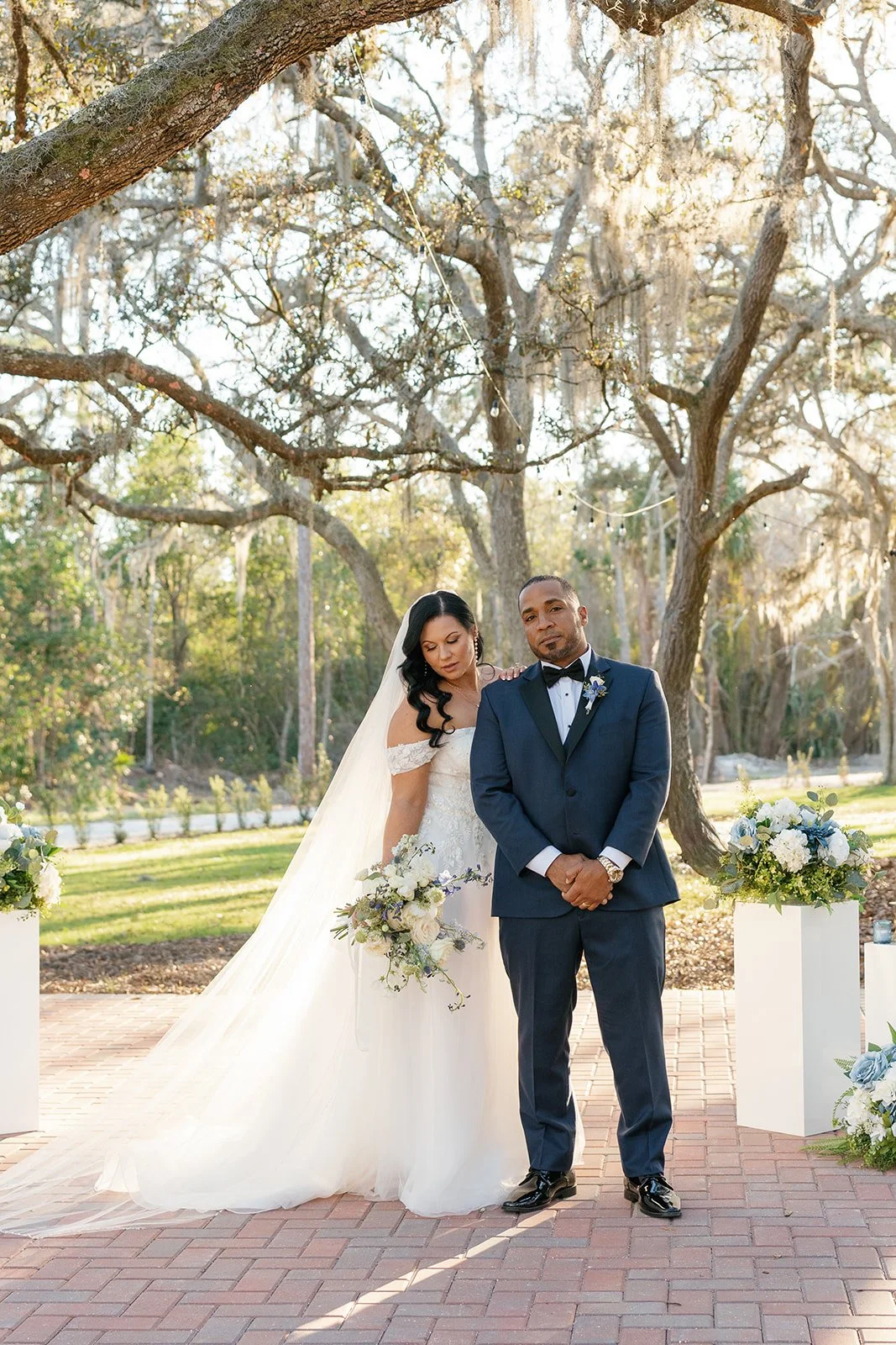 A bride and groom standing outdoors during a wedding ceremony. The bride is wearing a white wedding dress and veil, holding a bouquet of flowers. The groom is dressed in a navy blue tuxedo with a white shirt and black bow tie. They are under a large 