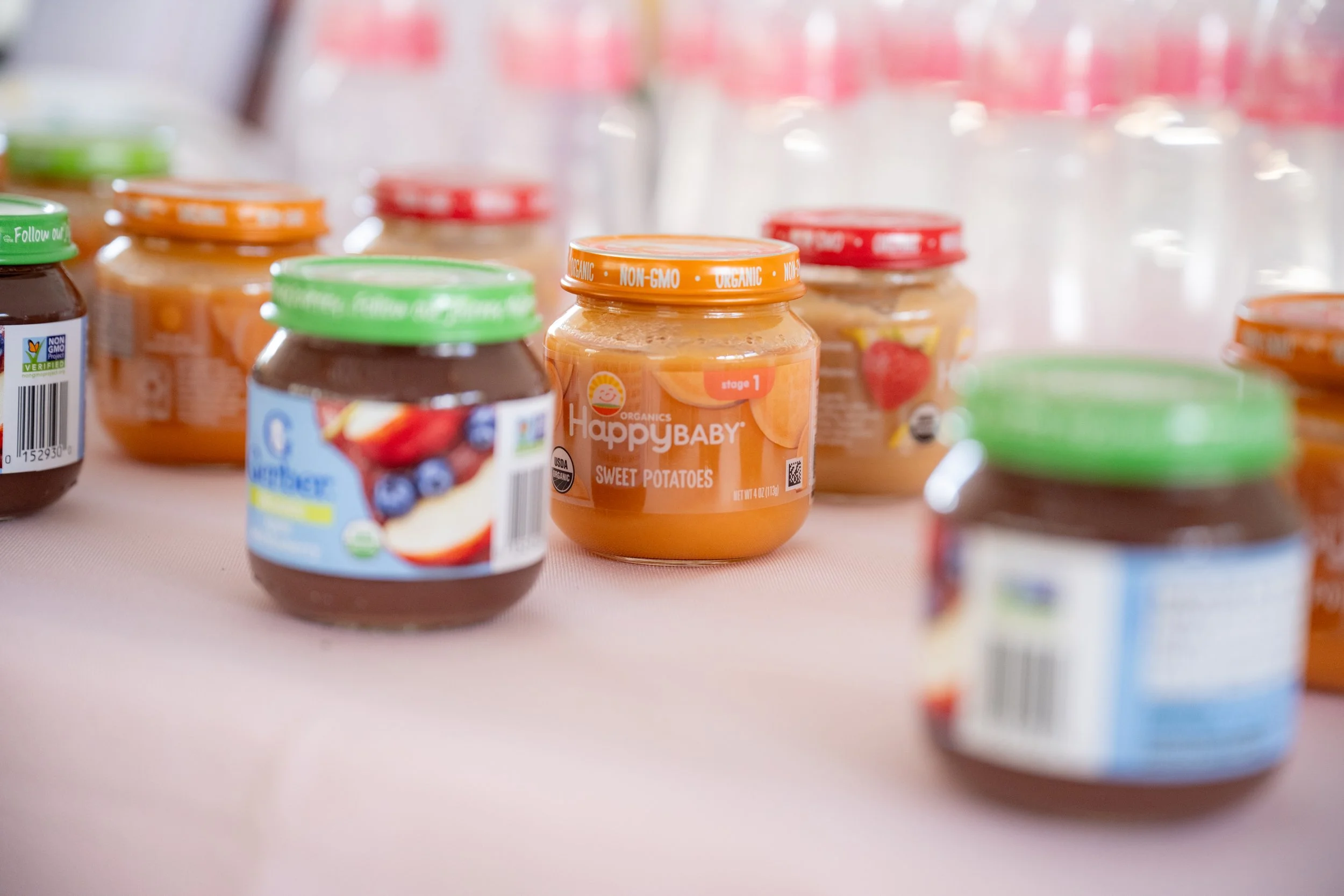 A close-up of multiple jars of organic baby food, including sweet potatoes, with colorful lids and labels, on a light-colored surface.