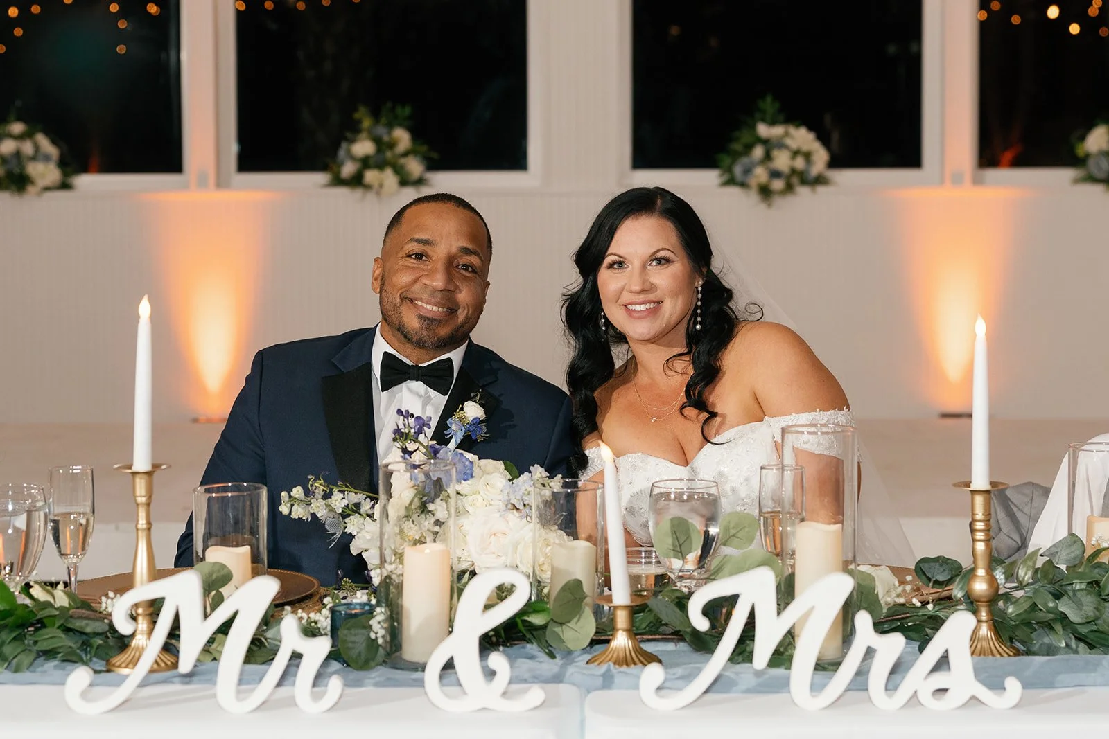Happy bride and groom sitting at wedding reception table with floral centerpiece, candles, and "Mr. & Mrs." sign, in front of large windows at night.