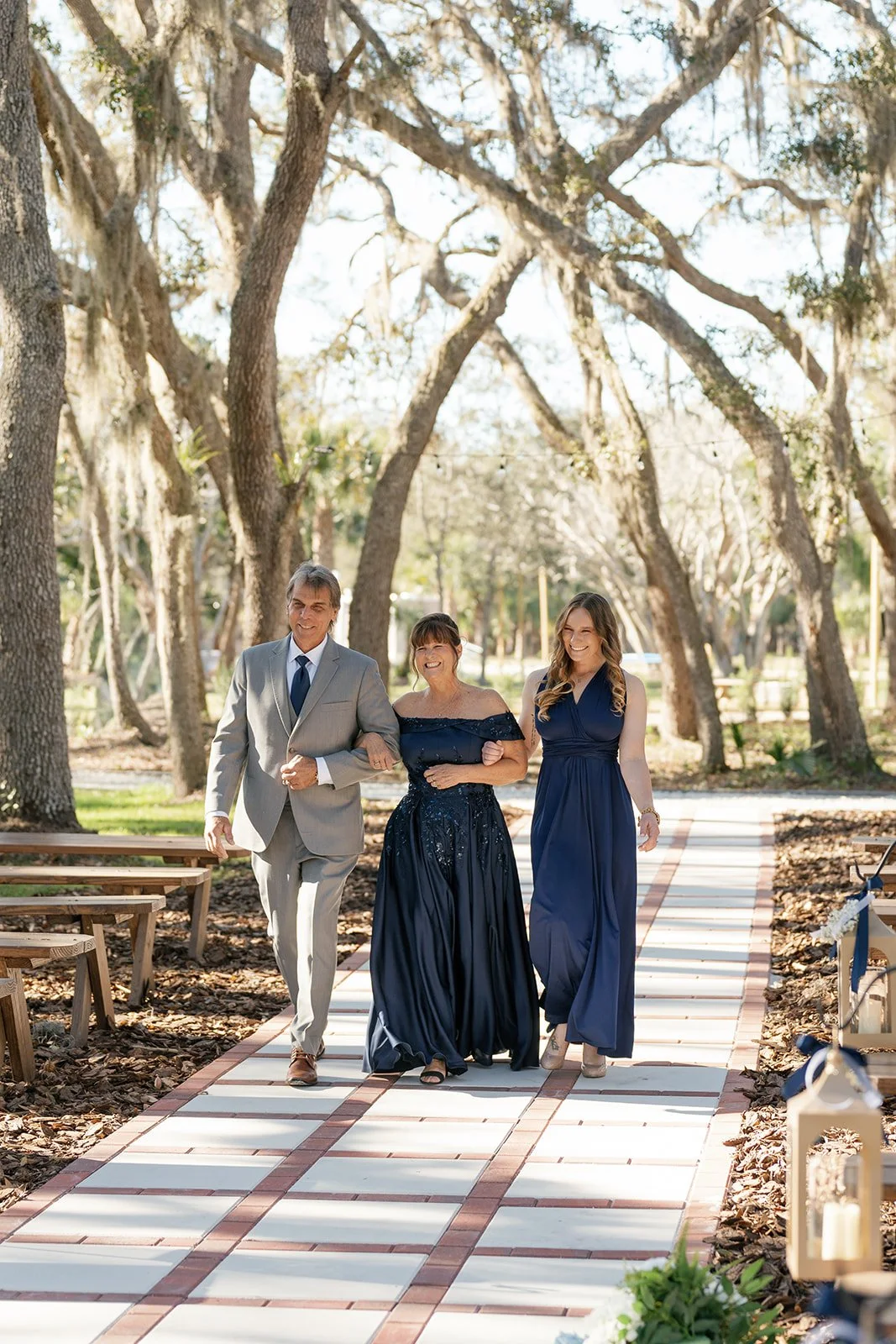 Three people walking arm in arm on a garden pathway during a sunny outdoor event, surrounded by tall trees with hanging moss.