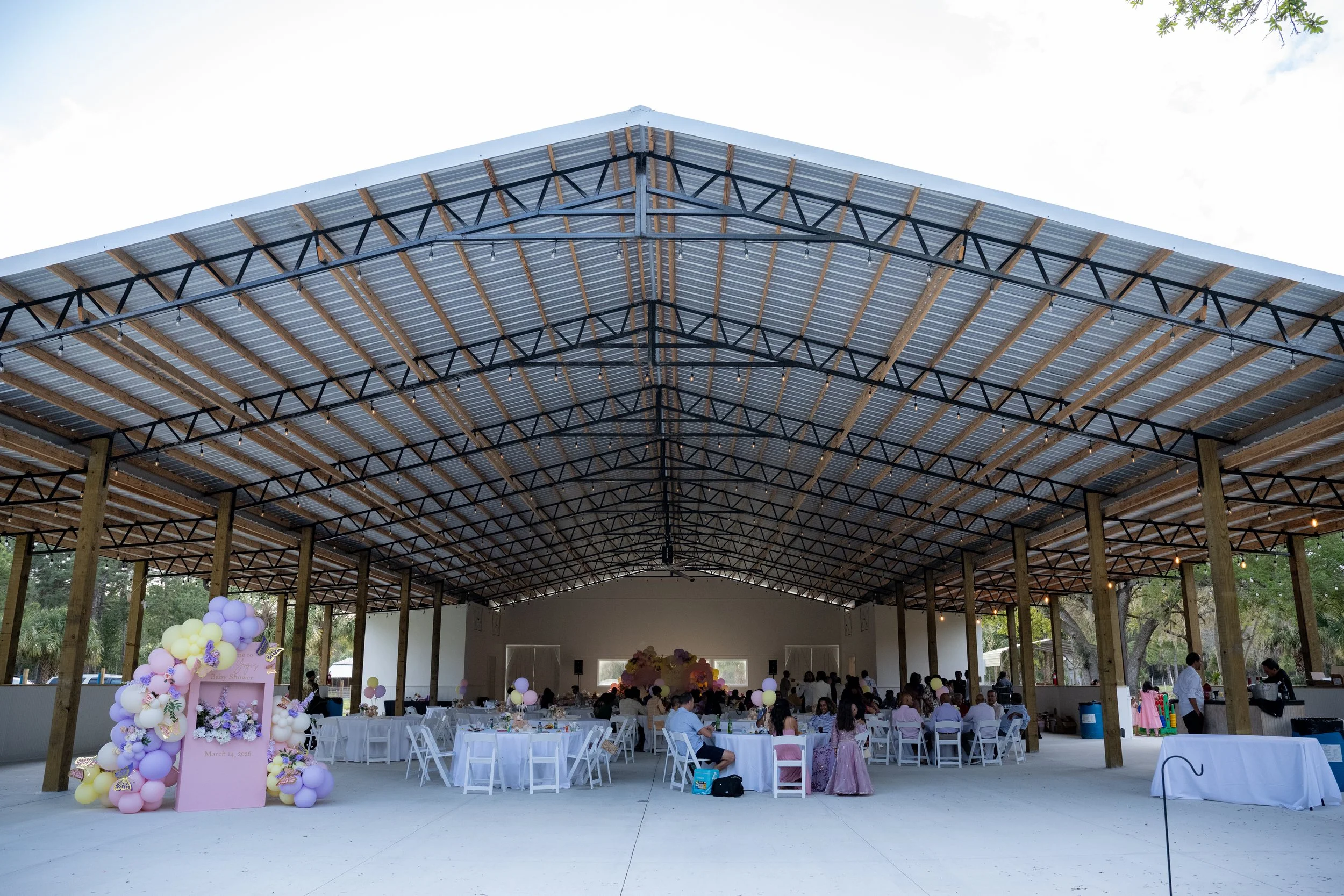 Indoor event space set up with decorated tables, pastel balloons, and guests, under a metal roof with wooden beams.