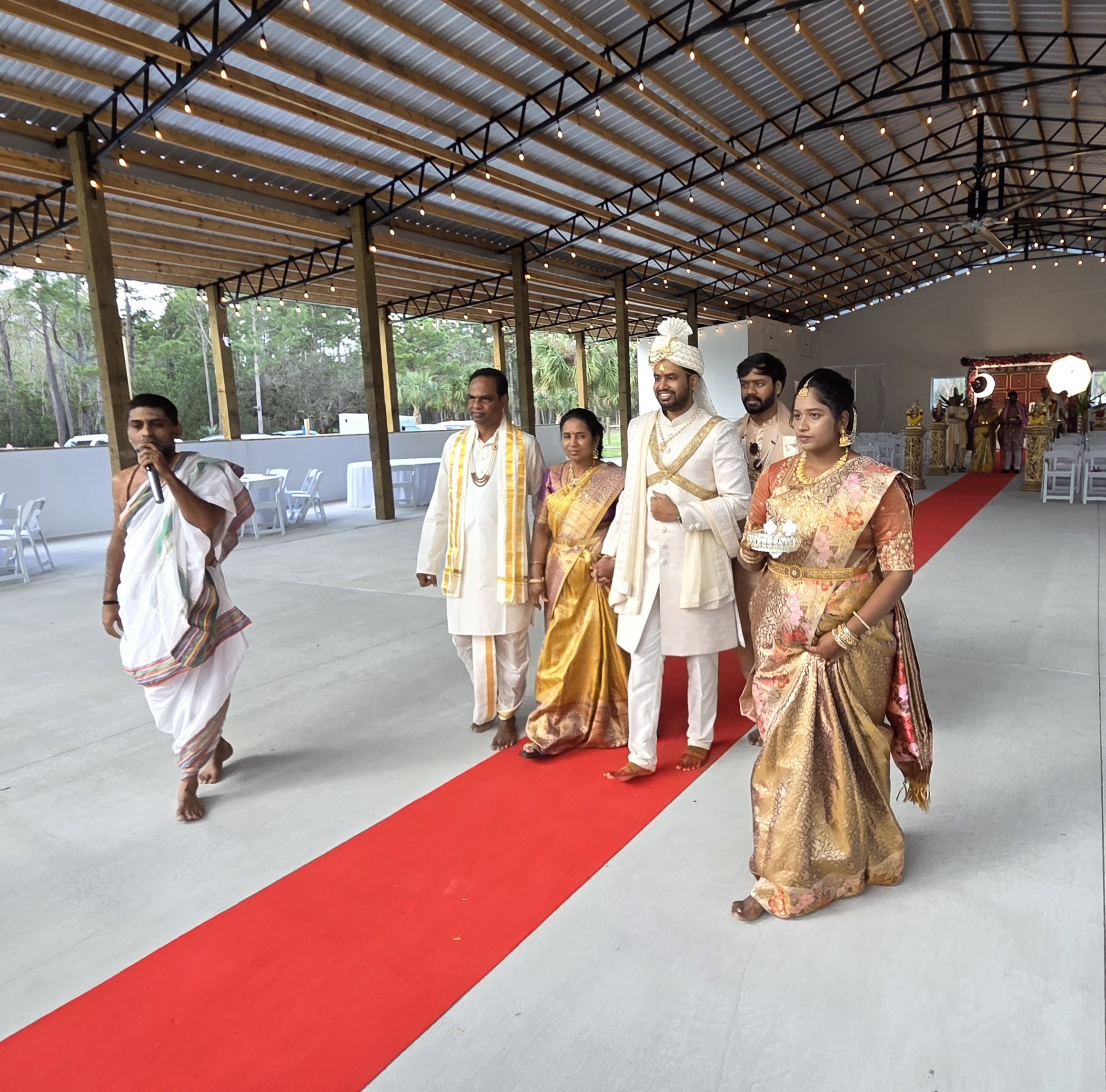 Indian wedding ceremony with people dressed in traditional attire standing on a red carpet under a metal and wood pavilion decorated with string lights.