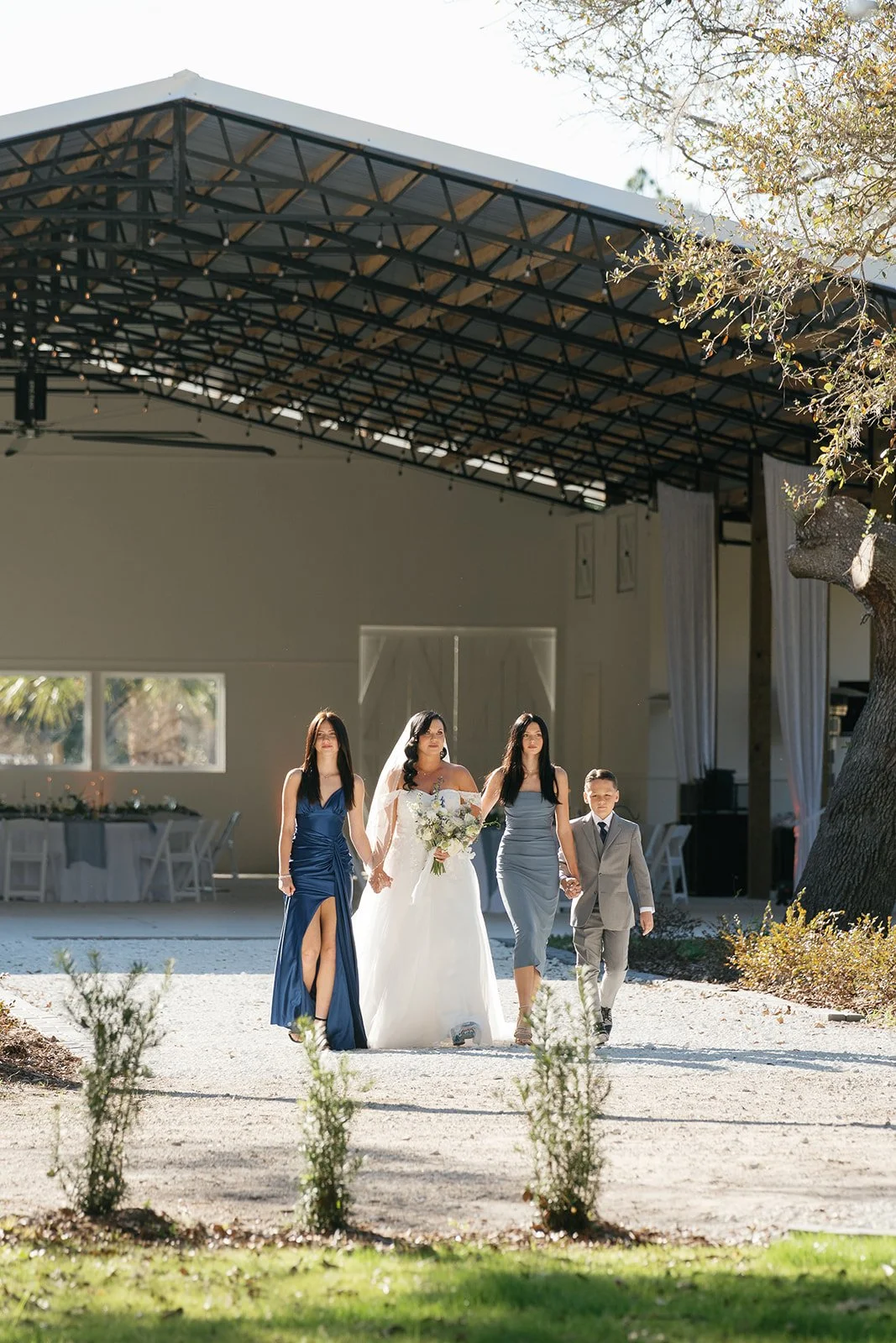 A bride in a white wedding dress holding a bouquet, accompanied by three young bridesmaids and a young boy, walking outdoors in front of a large open-air structure on a sunny day.