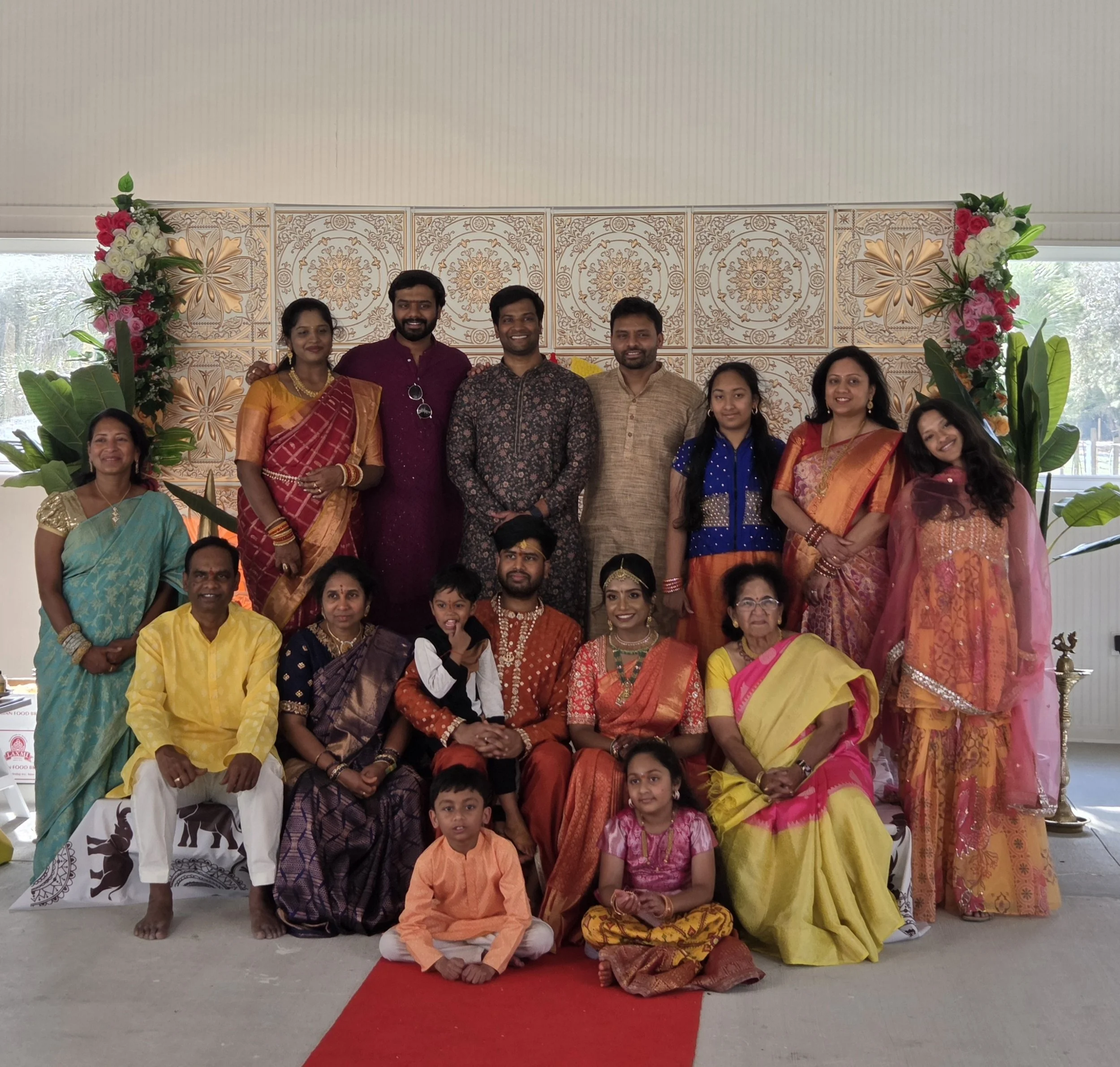 A group of men, women, and children dressed in colorful traditional Indian clothing, standing and sitting on a red carpet under a decorated arch with floral arrangements, posing for a photo in a bright indoor setting.