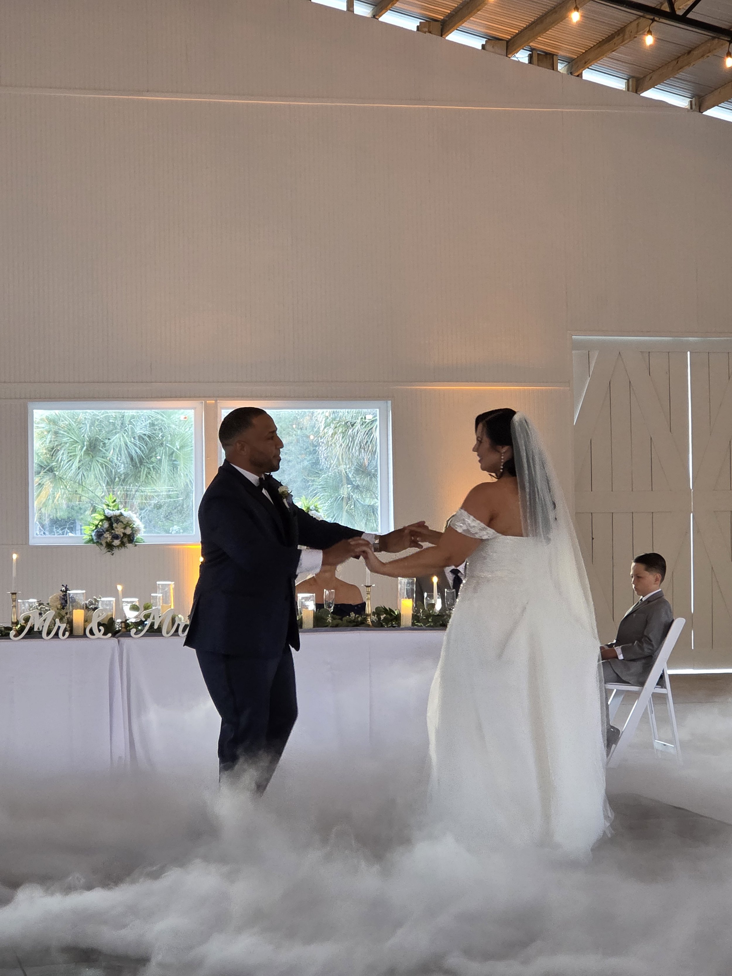 A bride and groom dancing at their wedding reception, with the bride in a white wedding dress and veil, and the groom in a dark tuxedo, in a decorated indoor venue with large windows and wedding table decor.