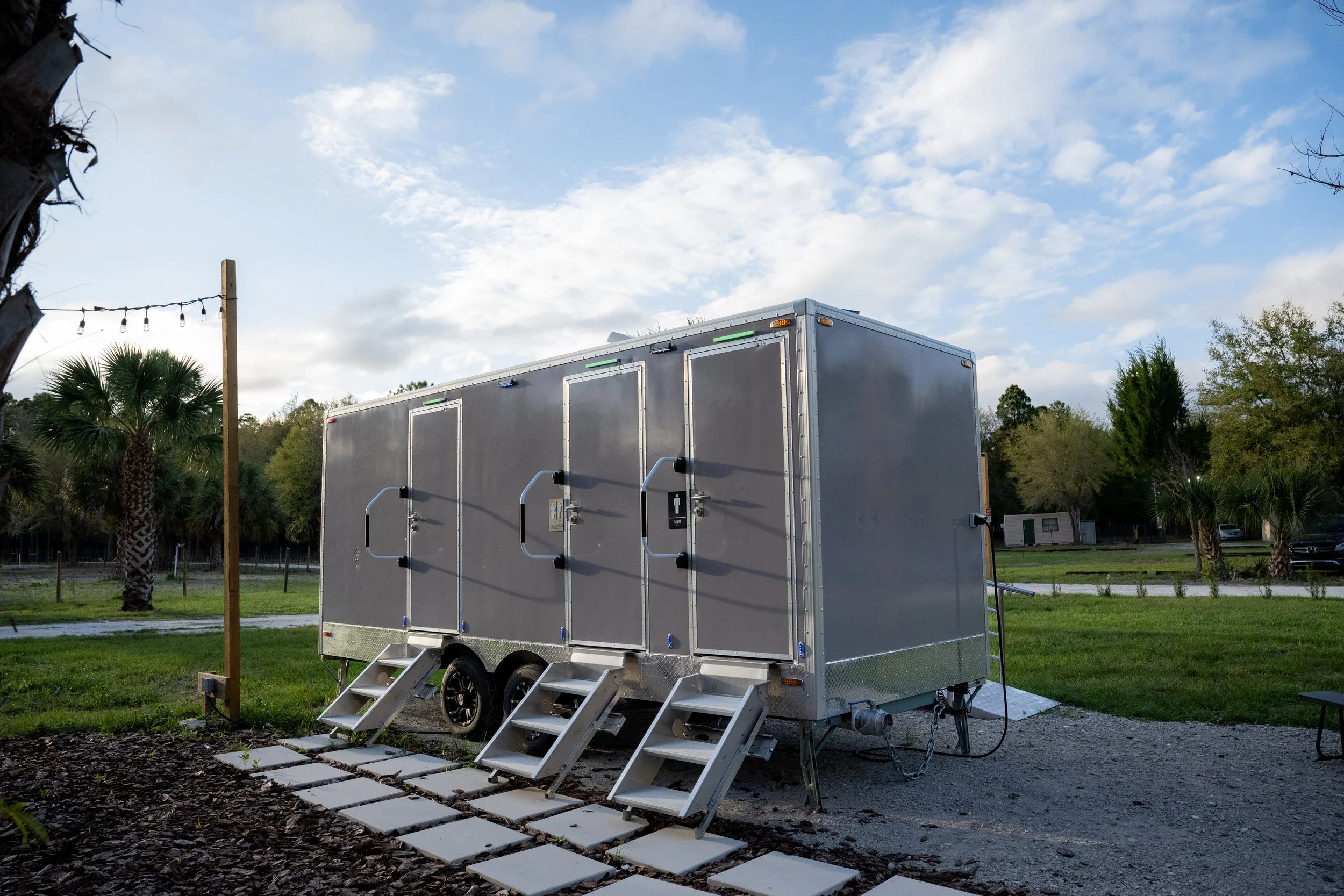 A mobile restroom trailer with steps leading up to its doors, situated on a grassy area with trees and a cloudy sky in the background.