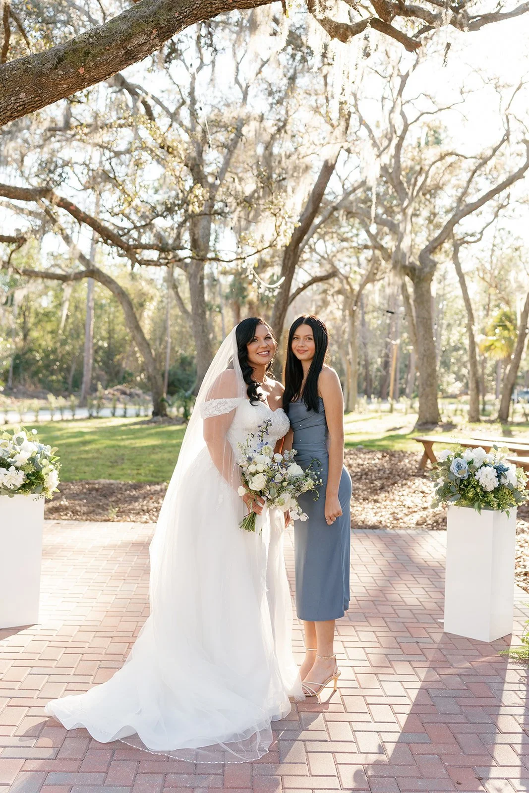 A bride in a white wedding gown holding a bouquet standing next to a woman in a blue dress, outdoors with trees and floral arrangements in the background.