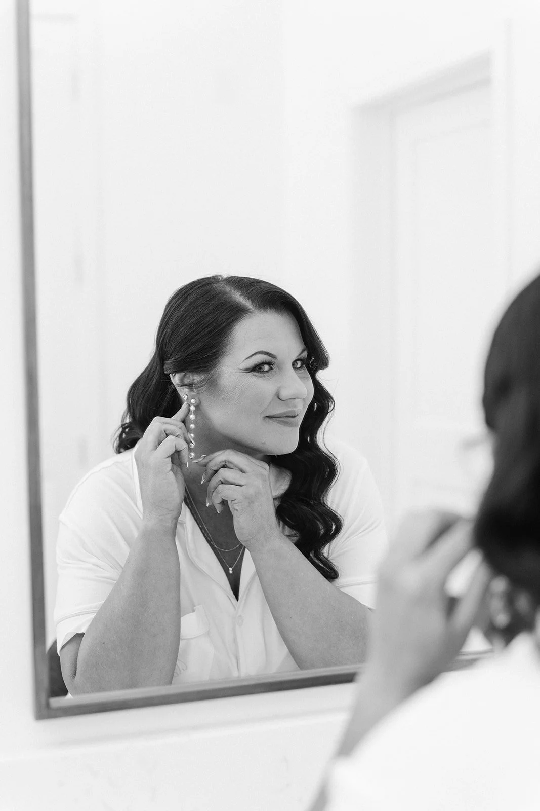 A woman with dark hair styled in loose waves looks at herself in a mirror, touching her earring, with a confident expression.