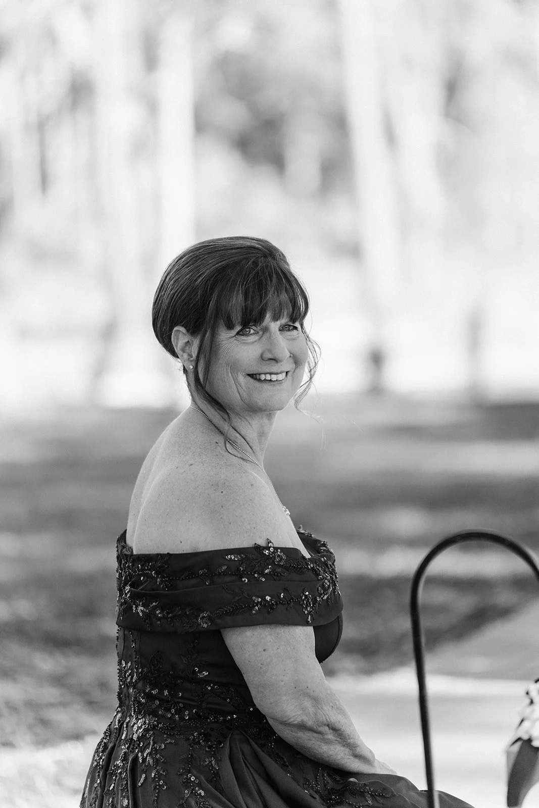 A smiling woman with dark, styled hair, wearing an off-the-shoulder dress with intricate beadwork, outdoors in black and white.
