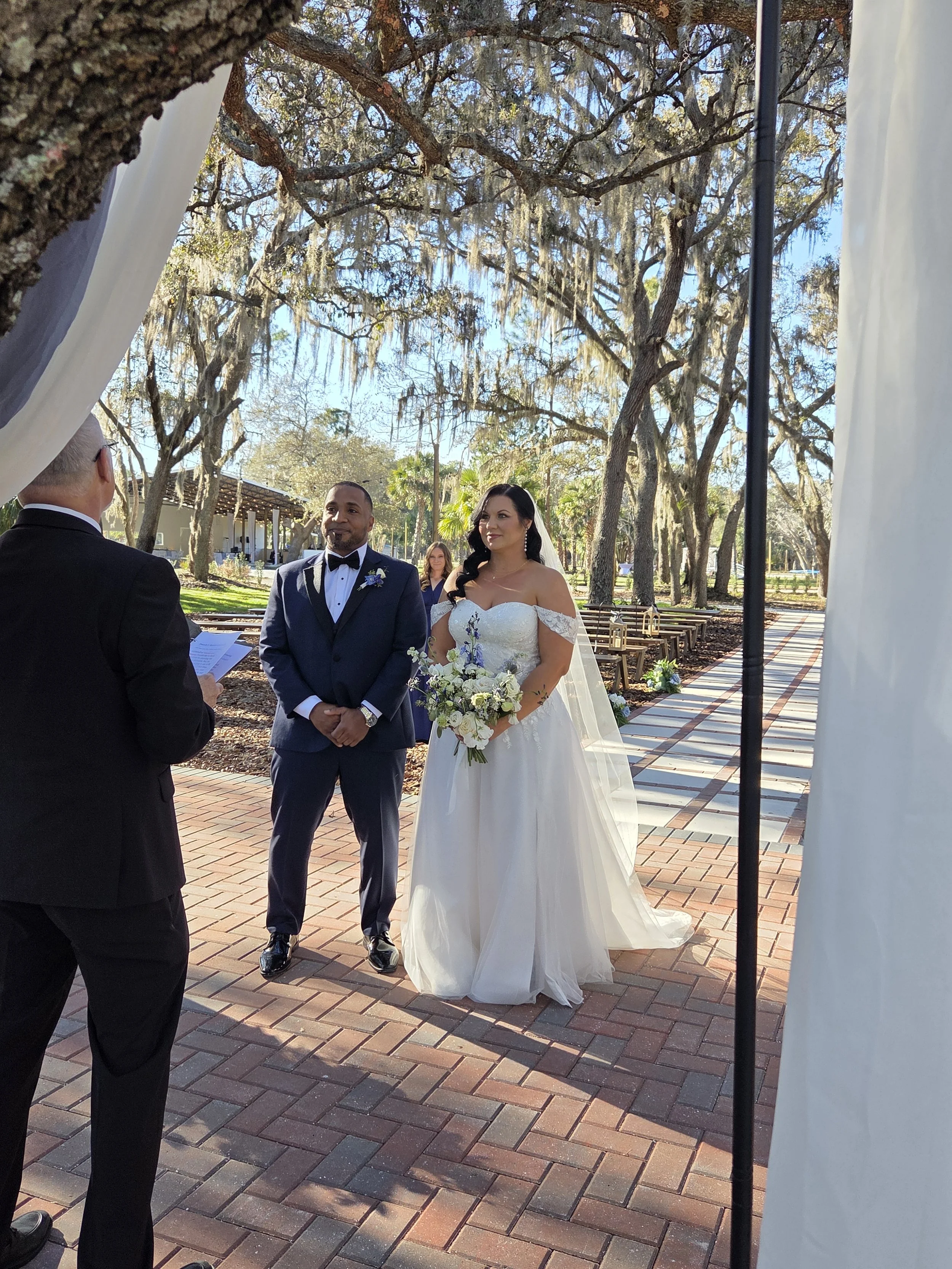 A bride and groom standing at the altar during their outdoor wedding ceremony, with a officiant on the left and a woman observer in the background, under a canopy of large trees with moss hanging from the branches.