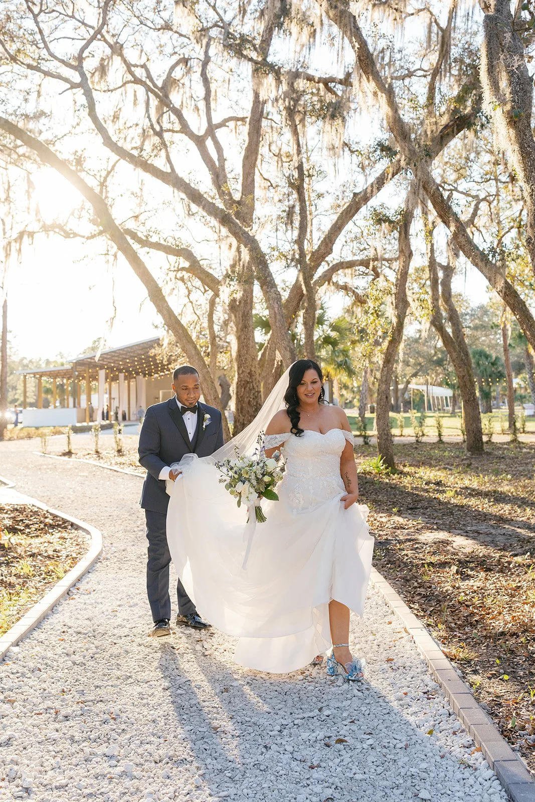 A bride and groom walking outdoors on a wedding day, with the bride holding a bouquet and wearing a white wedding dress, and the groom in a dark suit and bow tie, surrounded by trees and sunlight.