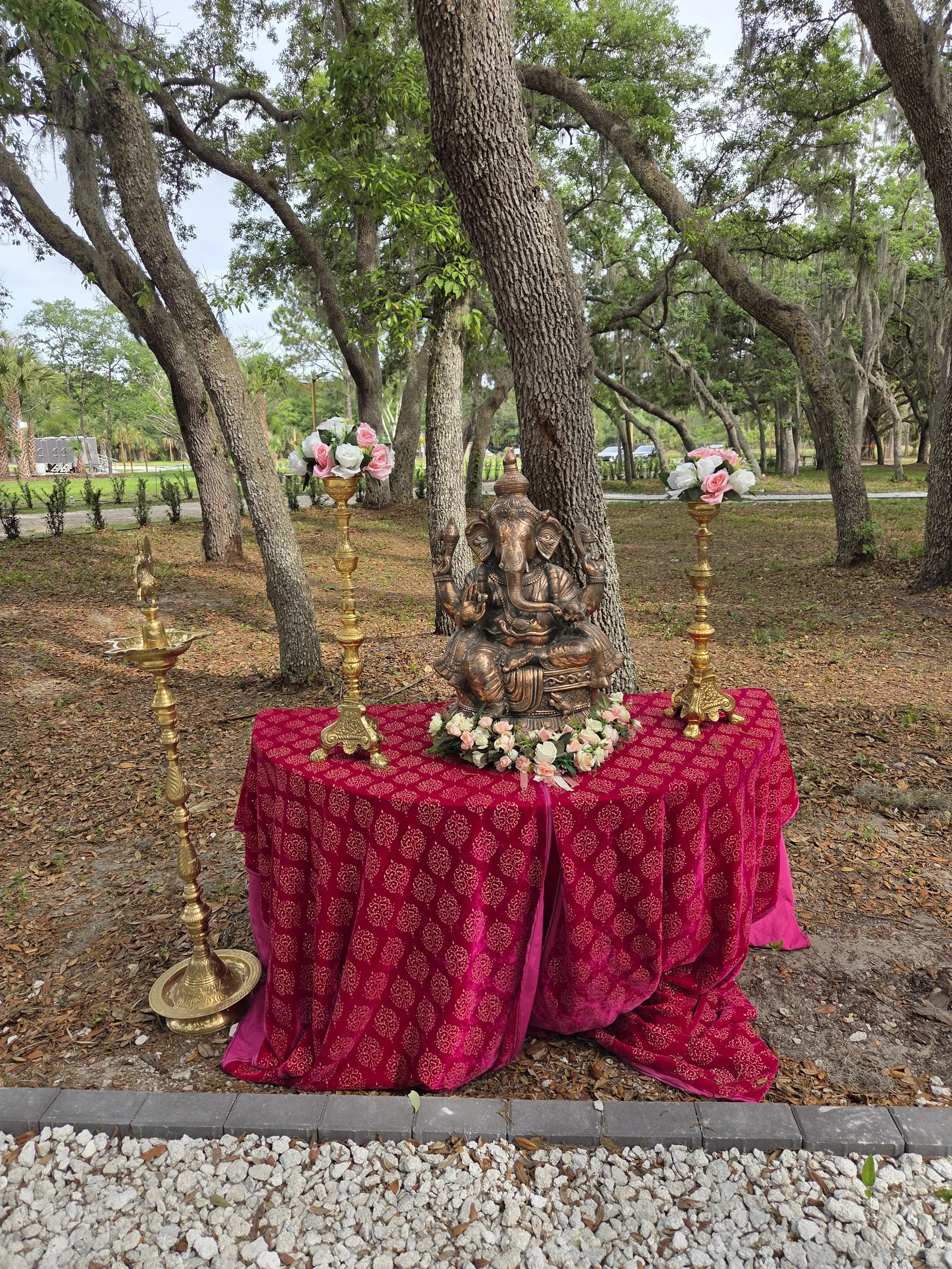 A brass table with a red cloth, decorated with a statue of Lord Ganesha, surrounded by flower arrangements, set outdoors among trees.