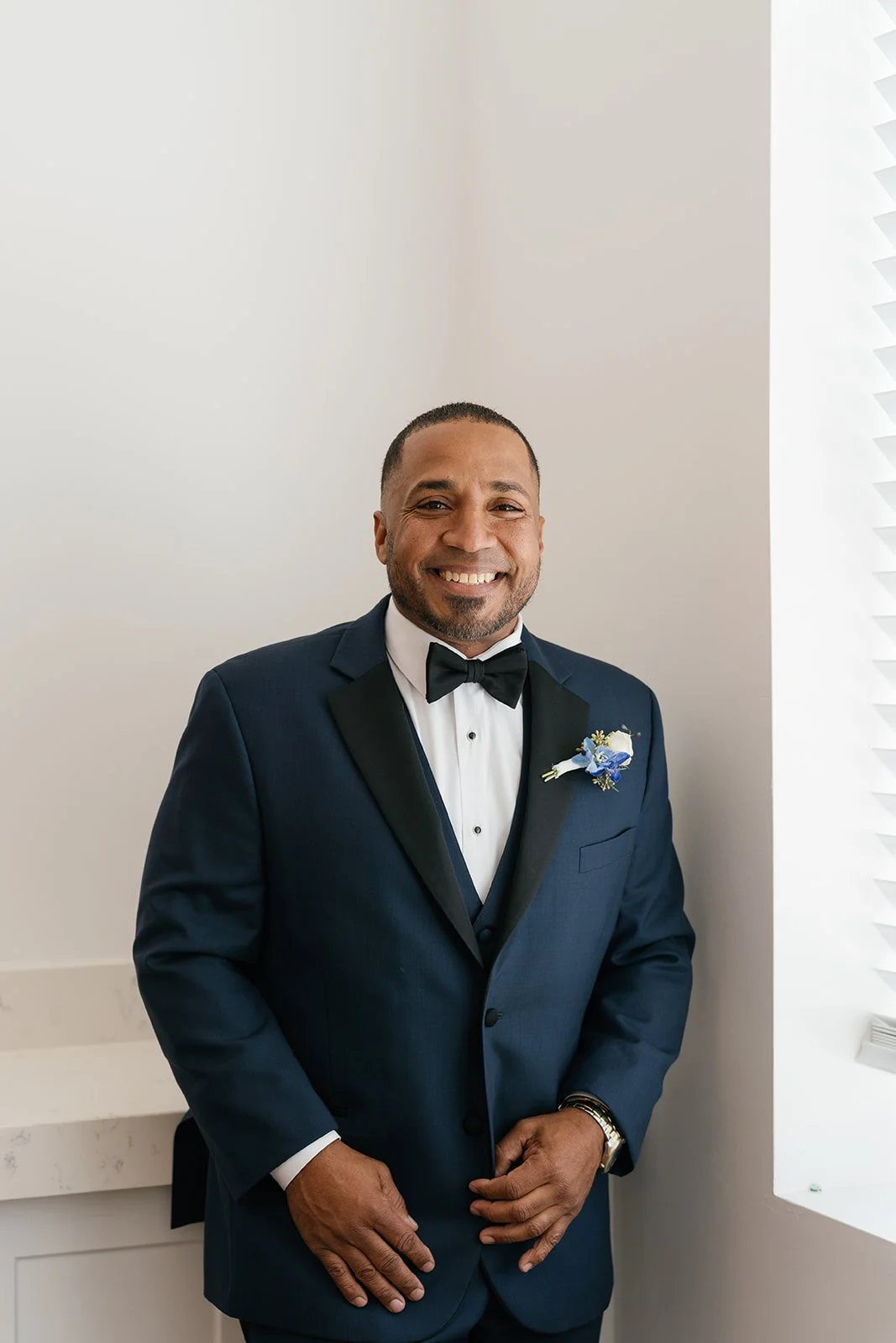 A man in a navy blue tuxedo with a black bow tie and white dress shirt, smiling and standing indoors near a white wall with blinds.