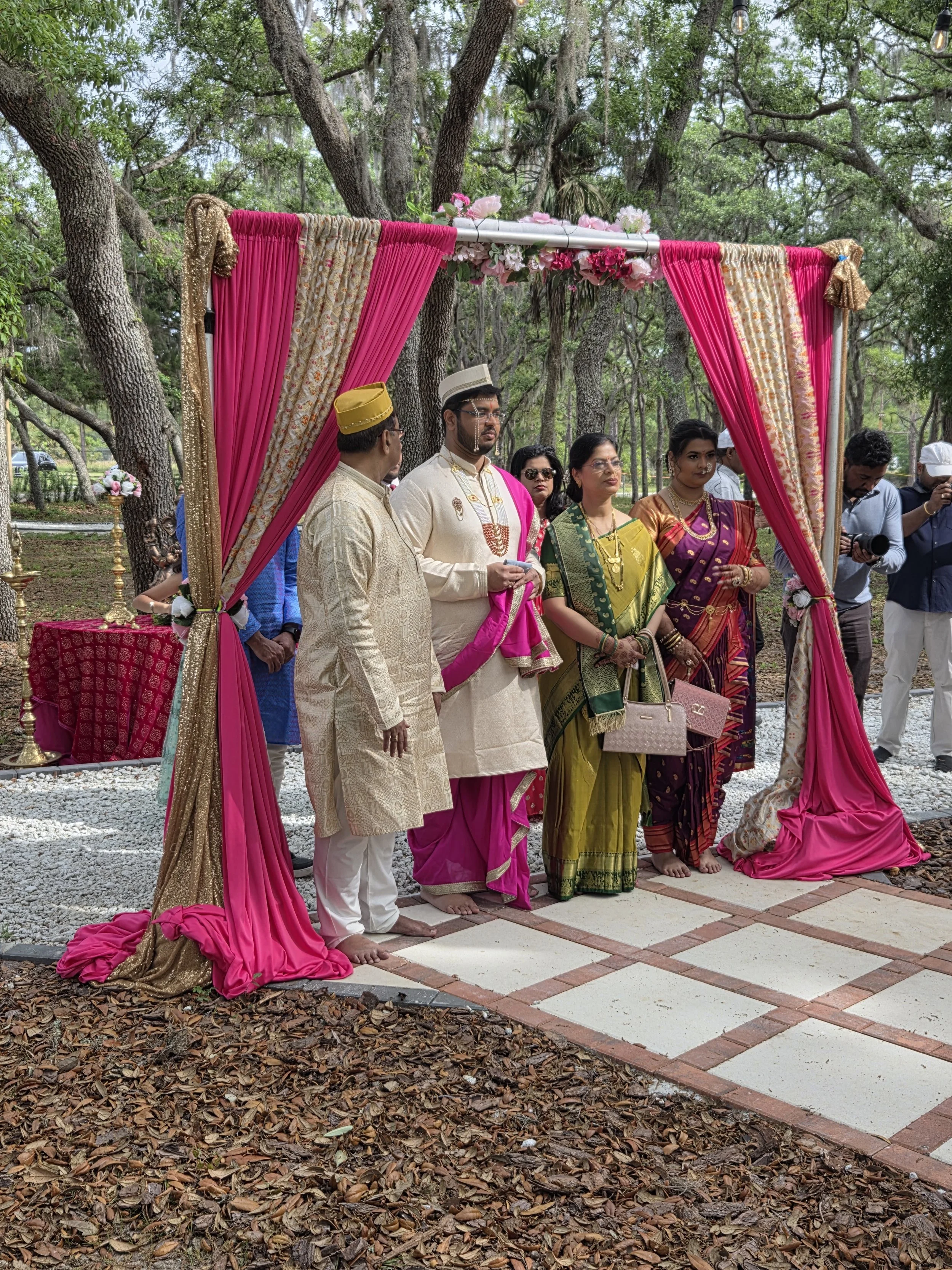 Group of people dressed in traditional Indian attire attending a wedding ceremony outdoors beneath a decorated canopy with pink and gold drapes, surrounded by trees and string lights.
