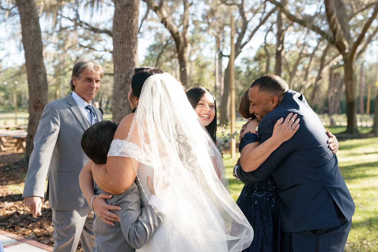 A wedding outdoor scene with people hugging and smiling, surrounded by trees.