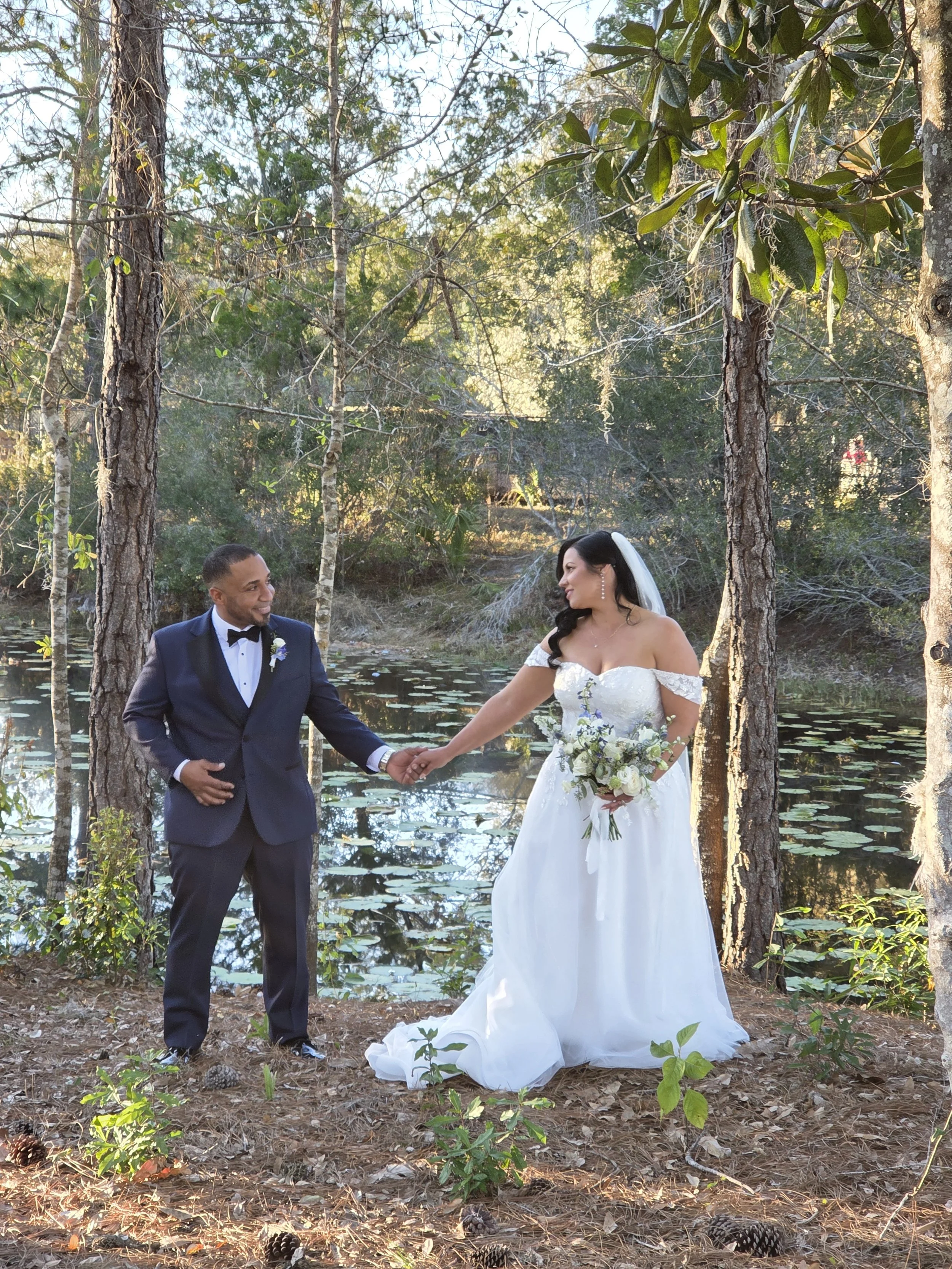 A bride and groom holding hands outdoors near a pond with trees and water lilies, surrounded by natural scenery.