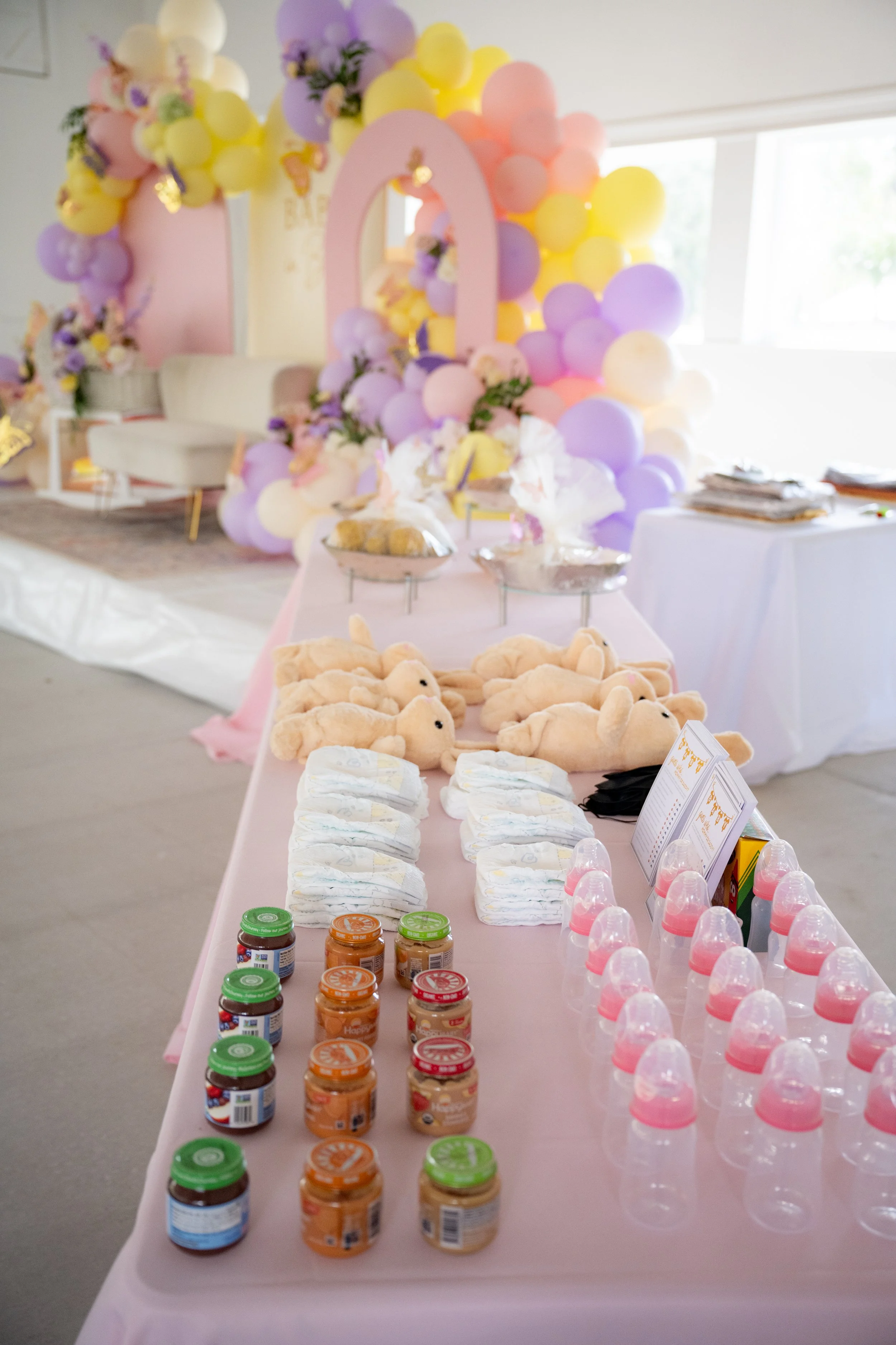 Decorations and food items for a baby shower, including a colorful balloon arch, plush teddy bears, baby bottles, jars of baby food, and diapers on a table.